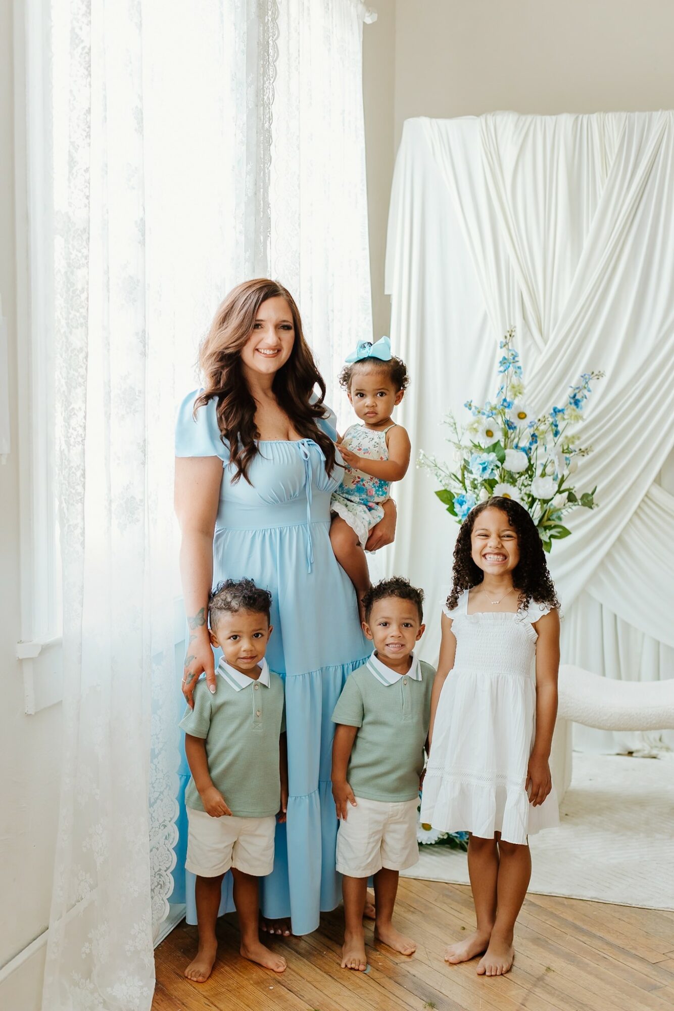 Woman with four children standing indoors near a window and floral arrangement, smiling, in light-colored clothing.