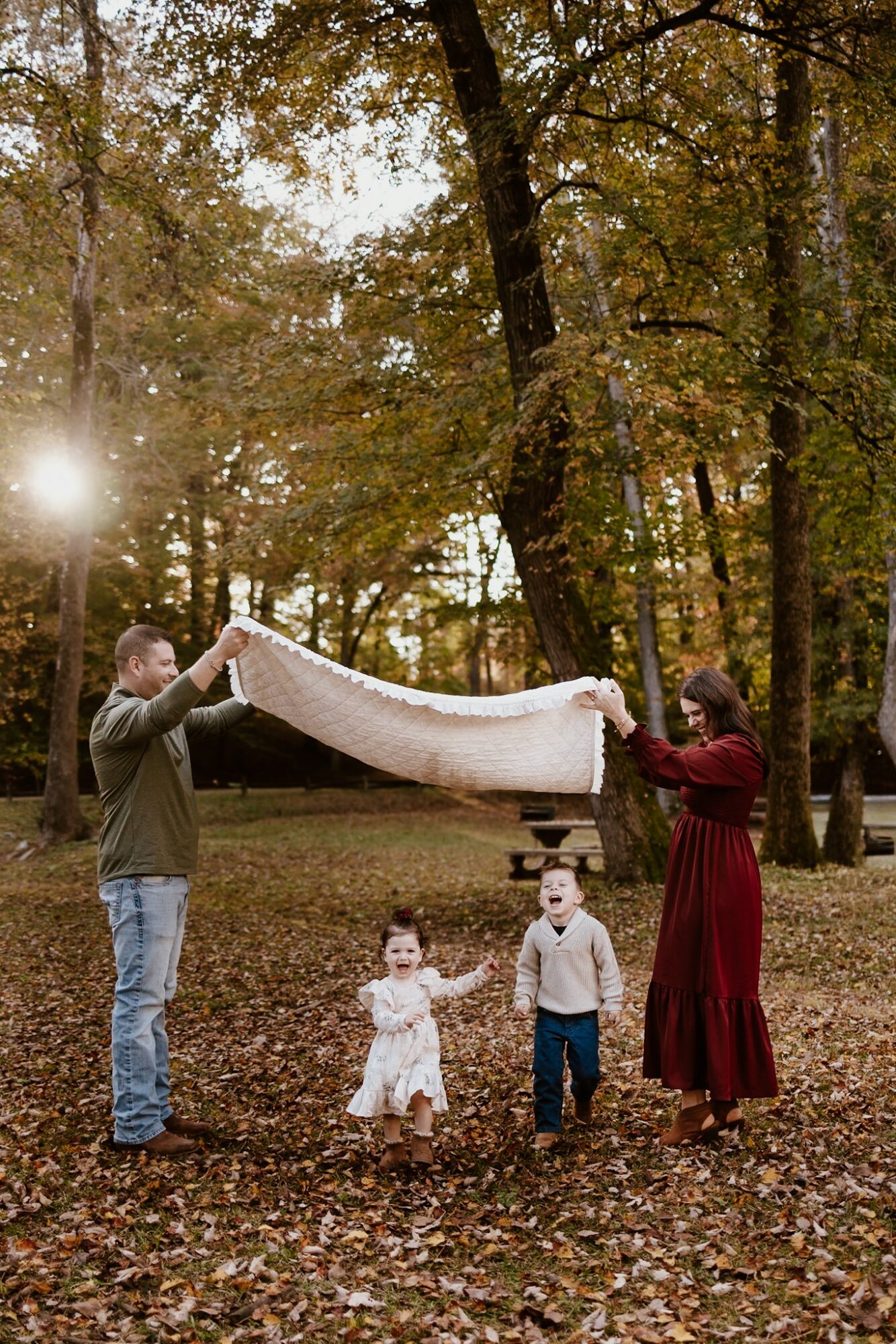 Family of four in a park with trees, holding a blanket, children standing on fallen leaves, sunlight filtering through trees.