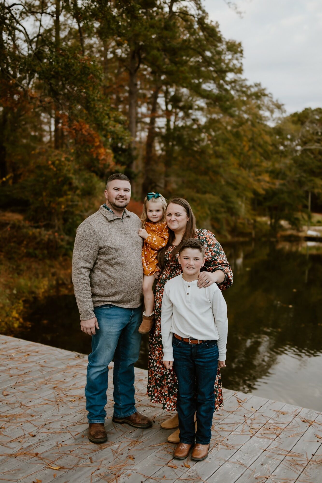 Family of four standing on a wooden dock near a lake with trees in autumn background, smiling at the camera.