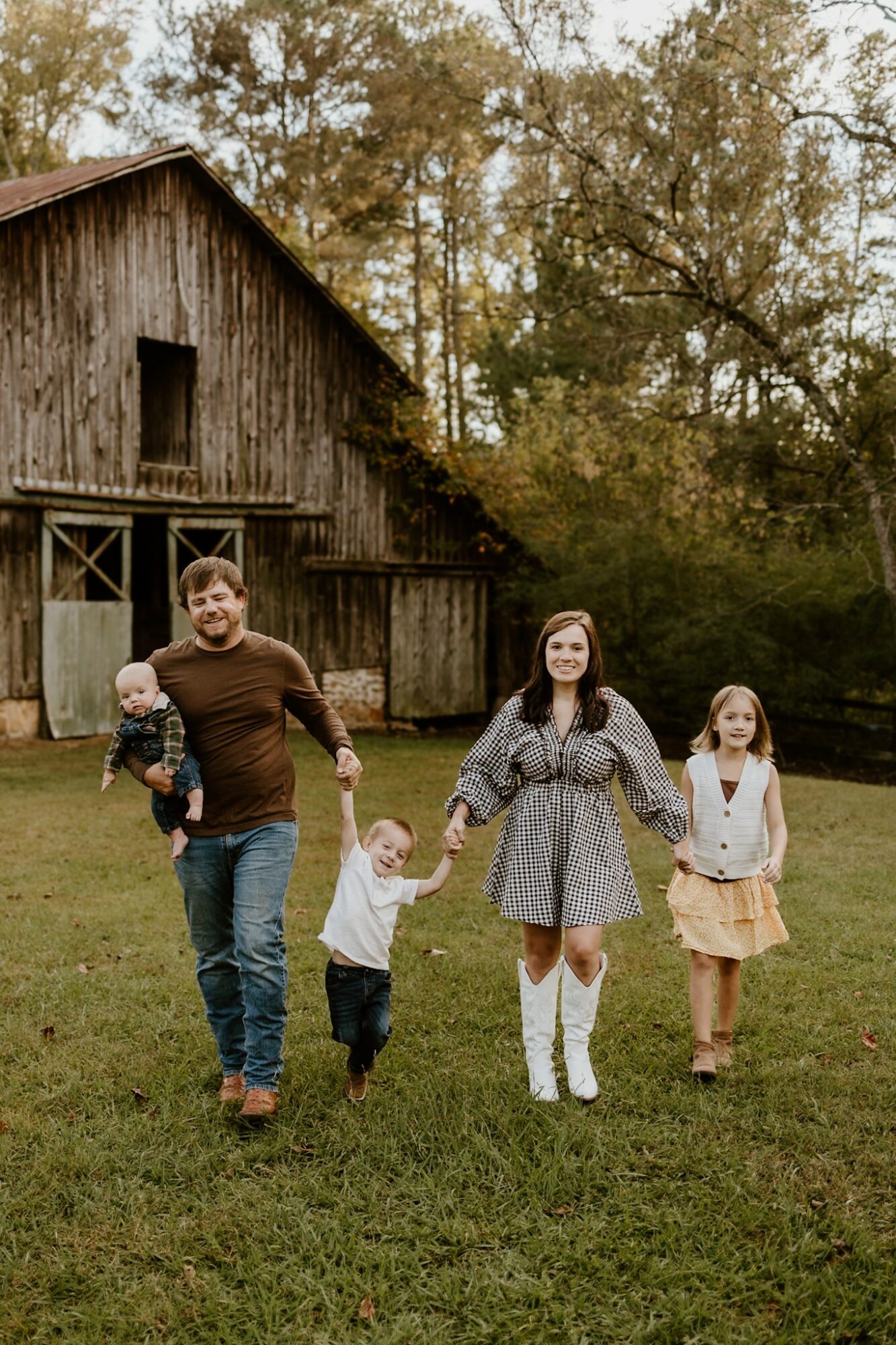 Family of five walking hand in hand outdoors near a wooden barn, trees in background, grassy field, autumn setting.