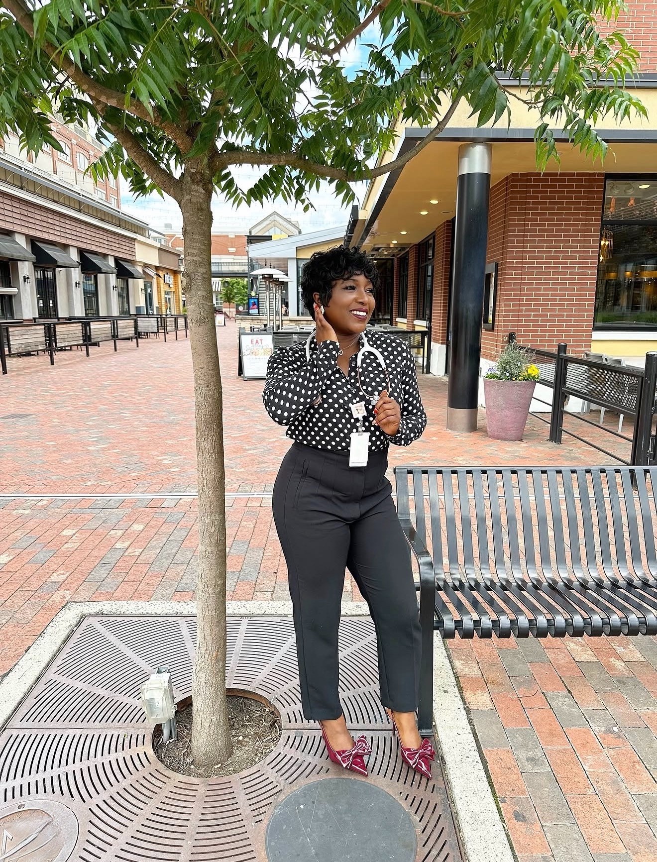 Woman standing next to a tree in an outdoor shopping area, smiling, wearing black pants, a polka dot blouse, and red shoes.