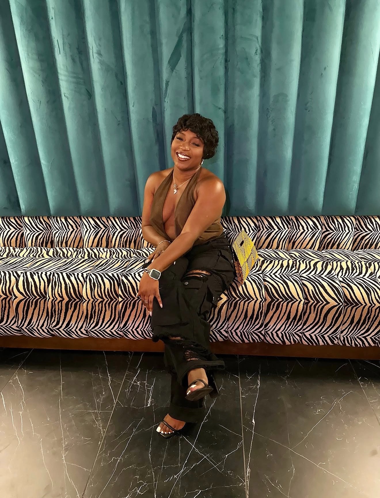 Smiling woman with short hair sitting on a patterned sofa in front of teal curtains, wearing a sleeveless top and black pants.
