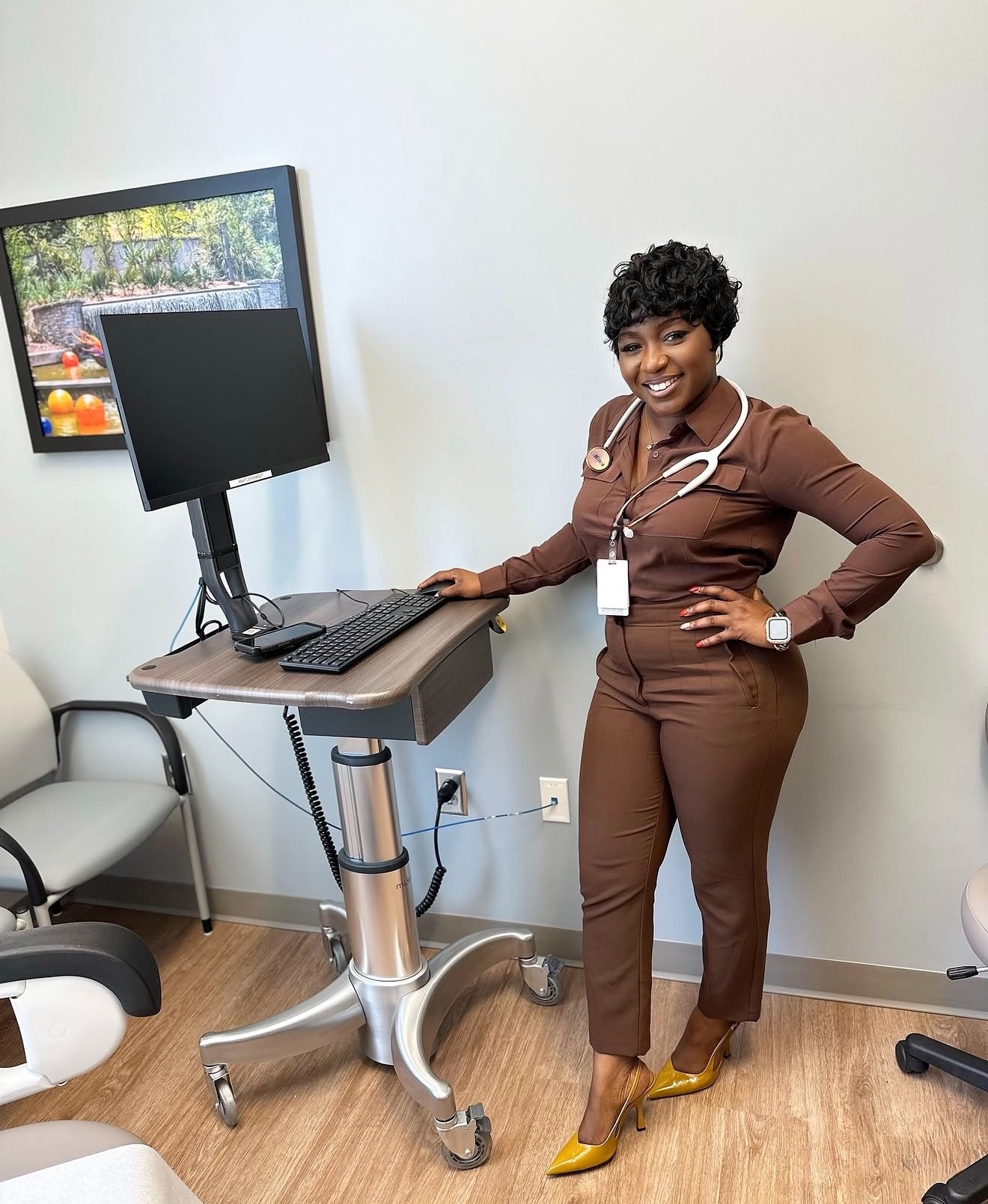Woman in brown professional attire standing in an office, smiling, with a computer on a desk nearby.