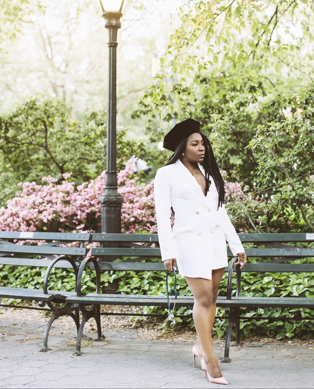 Woman in white blazer dress and black hat walking in park with greenery and pink flowers, holding a bag.