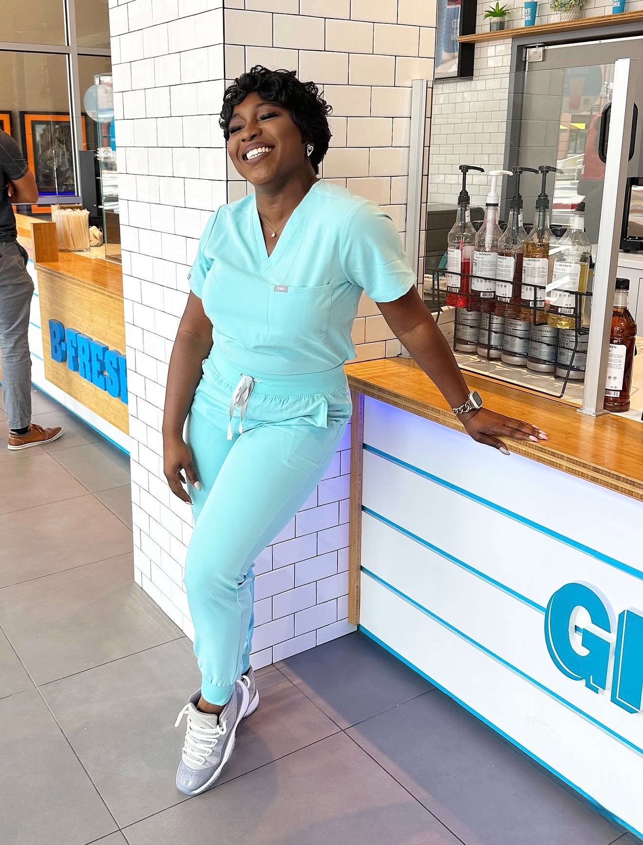 Smiling woman in light blue scrubs leaning on a counter in a modern shop or cafe, with a white brick wall behind her.