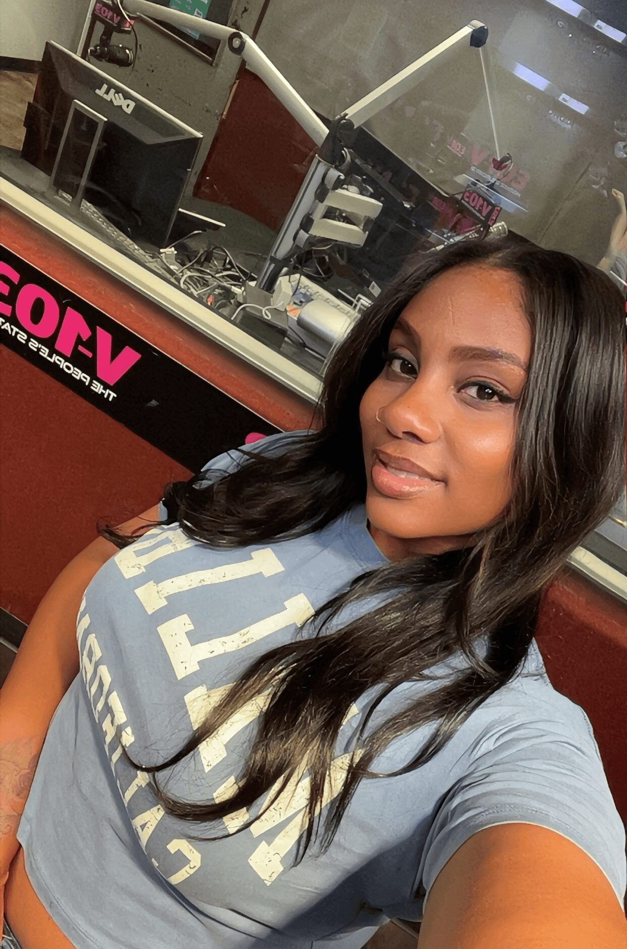 Young woman with long dark hair taking a selfie in a radio station studio, smiling slightly, wearing a light blue shirt.