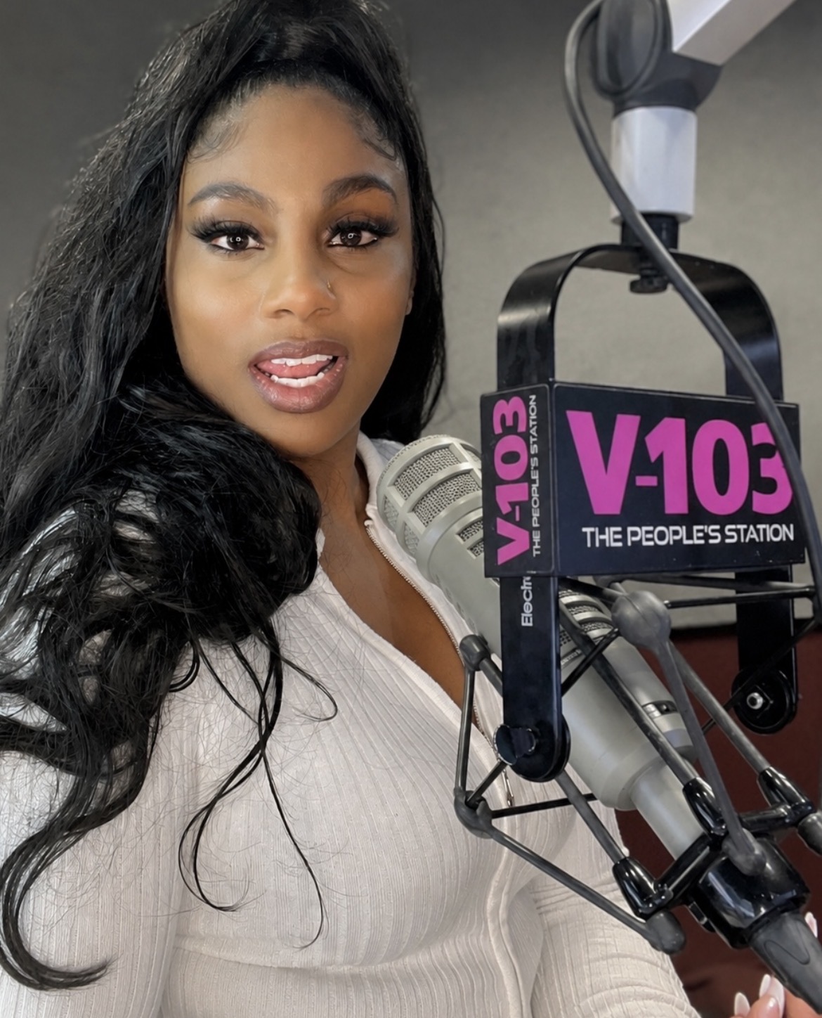Woman with long black hair speaking into a microphone at a radio station, V103 logo visible, wearing a white top.