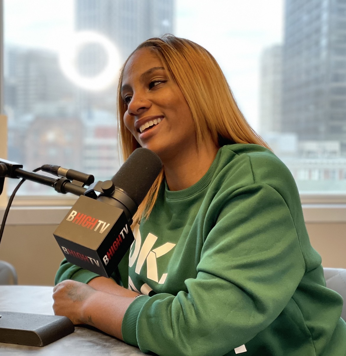Woman with long hair smiling, speaking into a microphone during a radio interview, wearing a green sweatshirt, cityscape background.