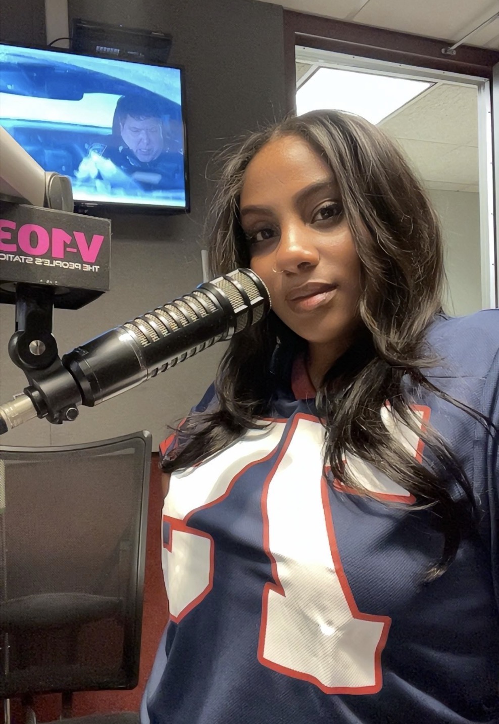 Woman with long dark hair speaking into a microphone in a radio studio, wearing a sports jersey, with a TV screen in the background.