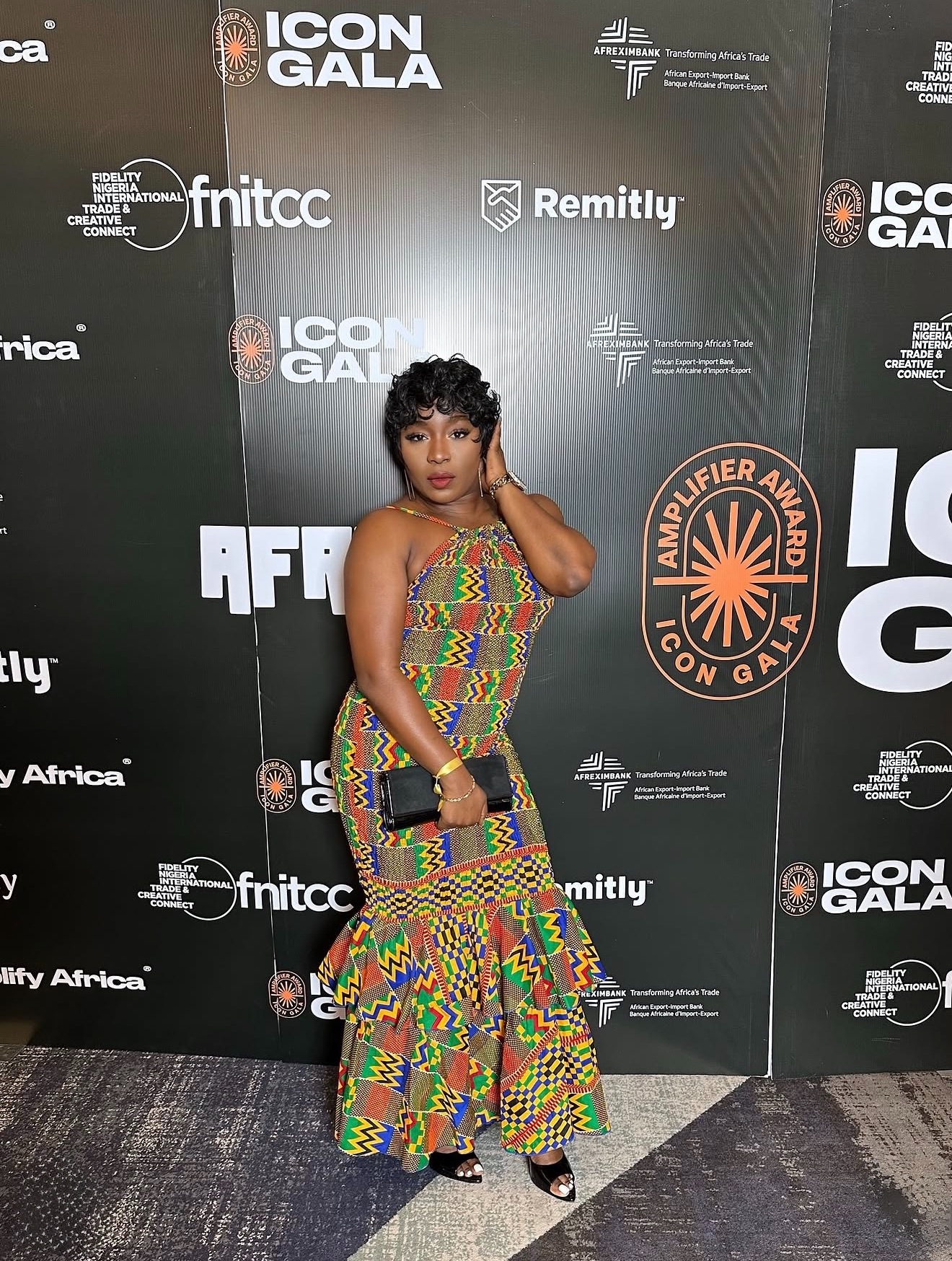 Woman in colorful patterned dress holding a clutch, standing on a black carpeted floor in front of a black backdrop with logos.