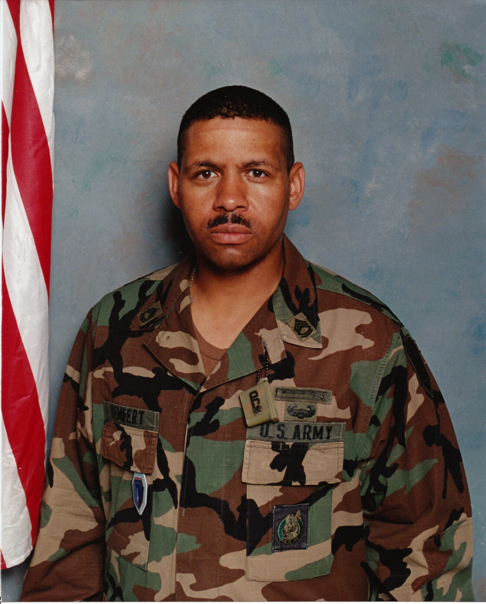 Man in U.S. Army camouflage uniform standing in front of a blue background with an American flag partially visible on the left.