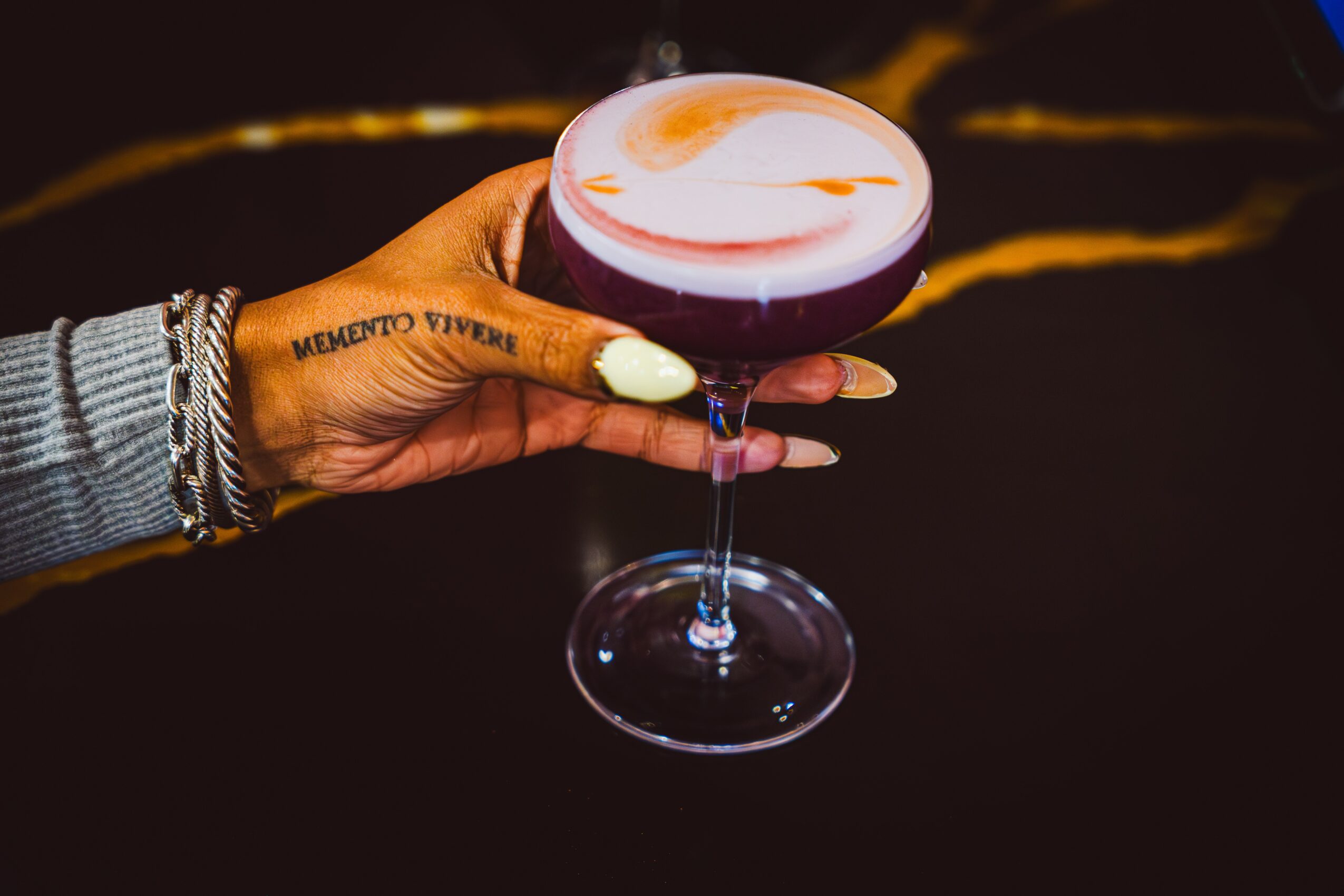 Hand holding a cocktail glass with a layered drink, against a dark background. Person wears a gray sleeve and jewelry.