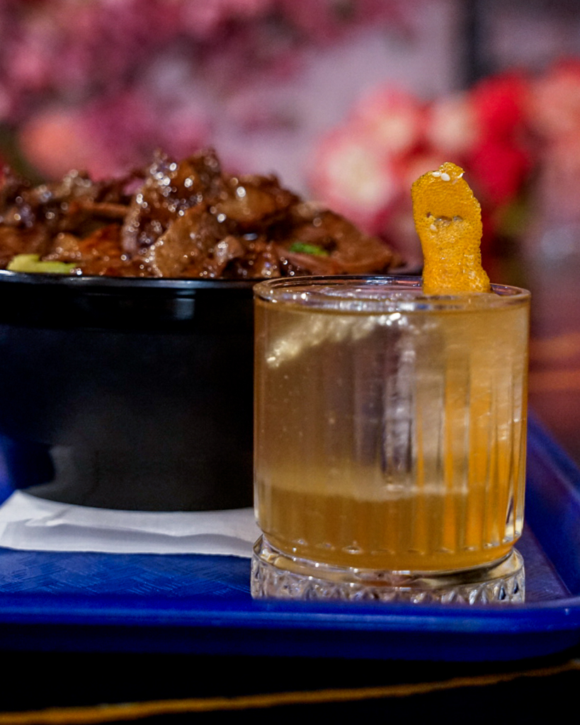 A glass of amber-colored drink with a lemon twist garnish, placed on a blue tray with food in the background.