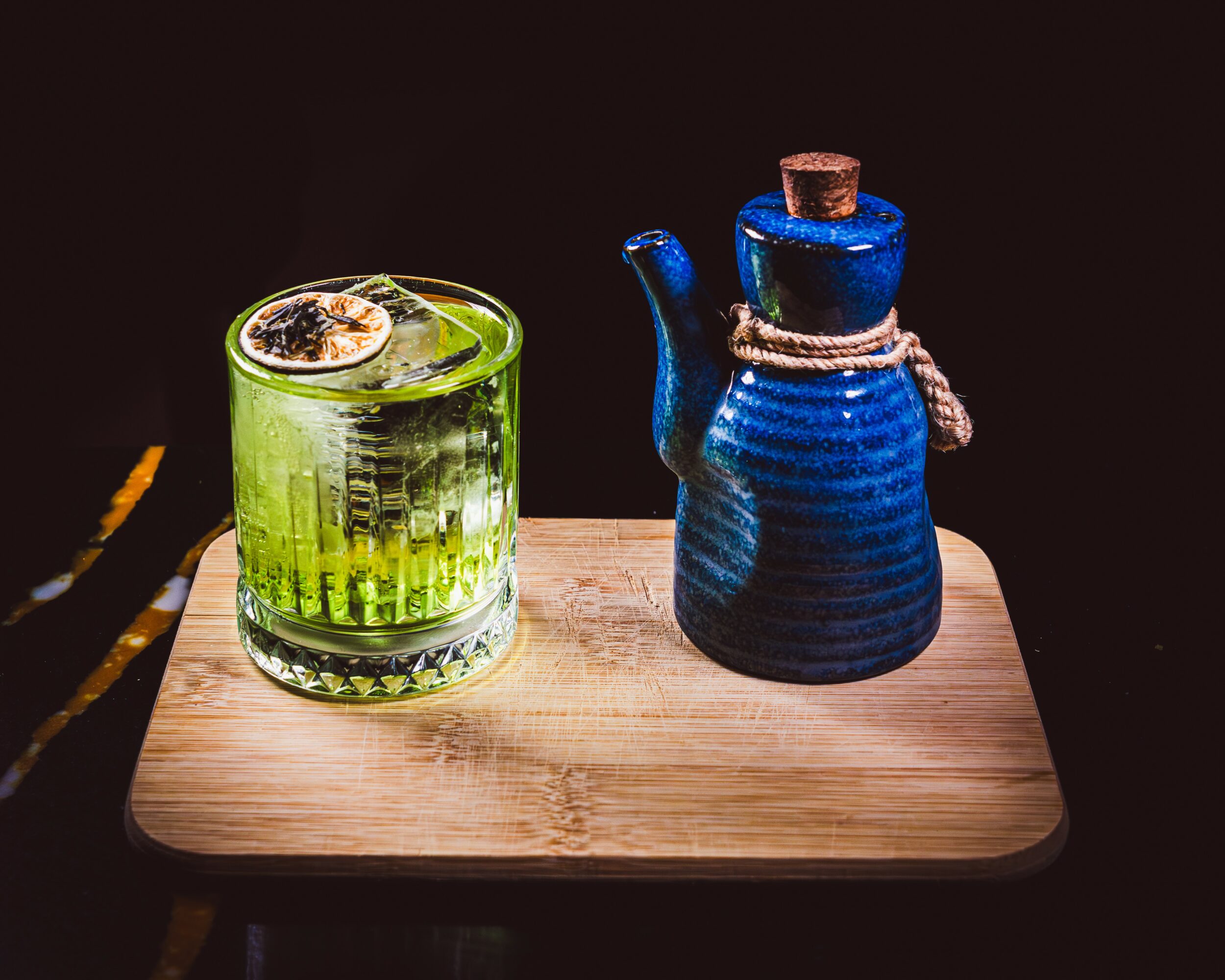 A green drink with a lemon slice in a glass and a blue ceramic teapot on a wooden tray.