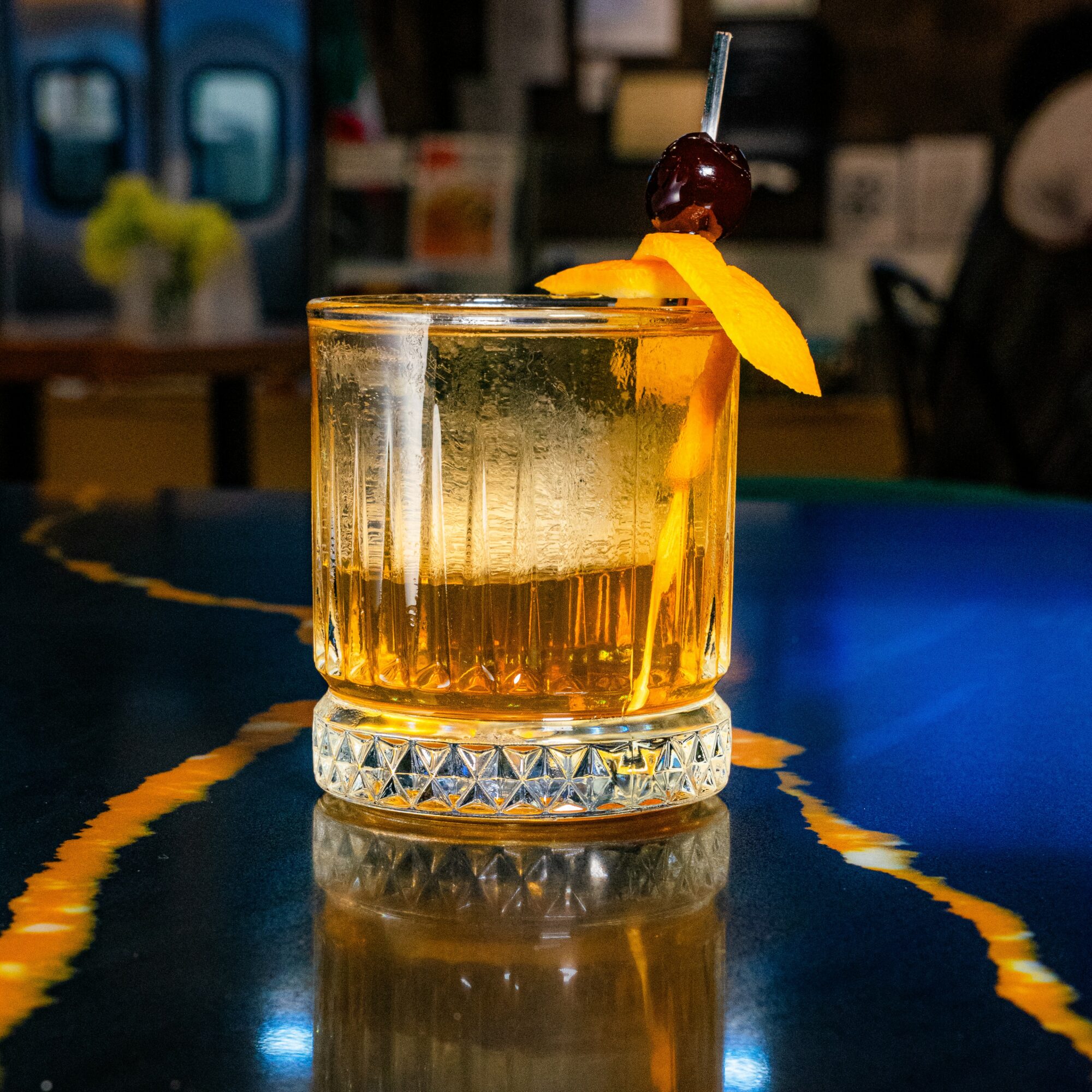 A glass of amber-colored drink with a cherry and orange peel garnish on a reflective surface.