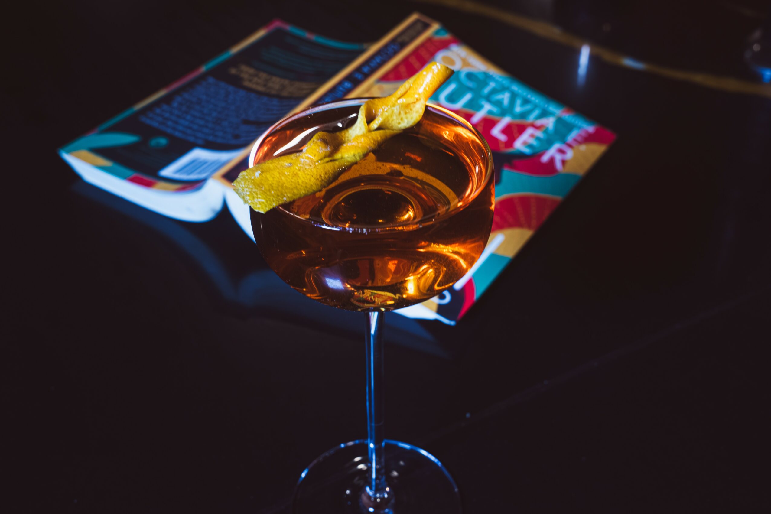 Cocktail glass with lemon peel and cherry, magazine in background on dark surface.