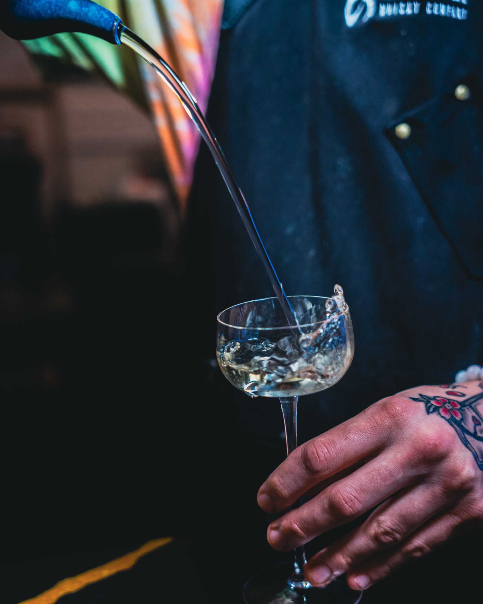 Person pouring clear liquid into a glass, with tattoos visible on hand, dark background, and colorful objects nearby.
