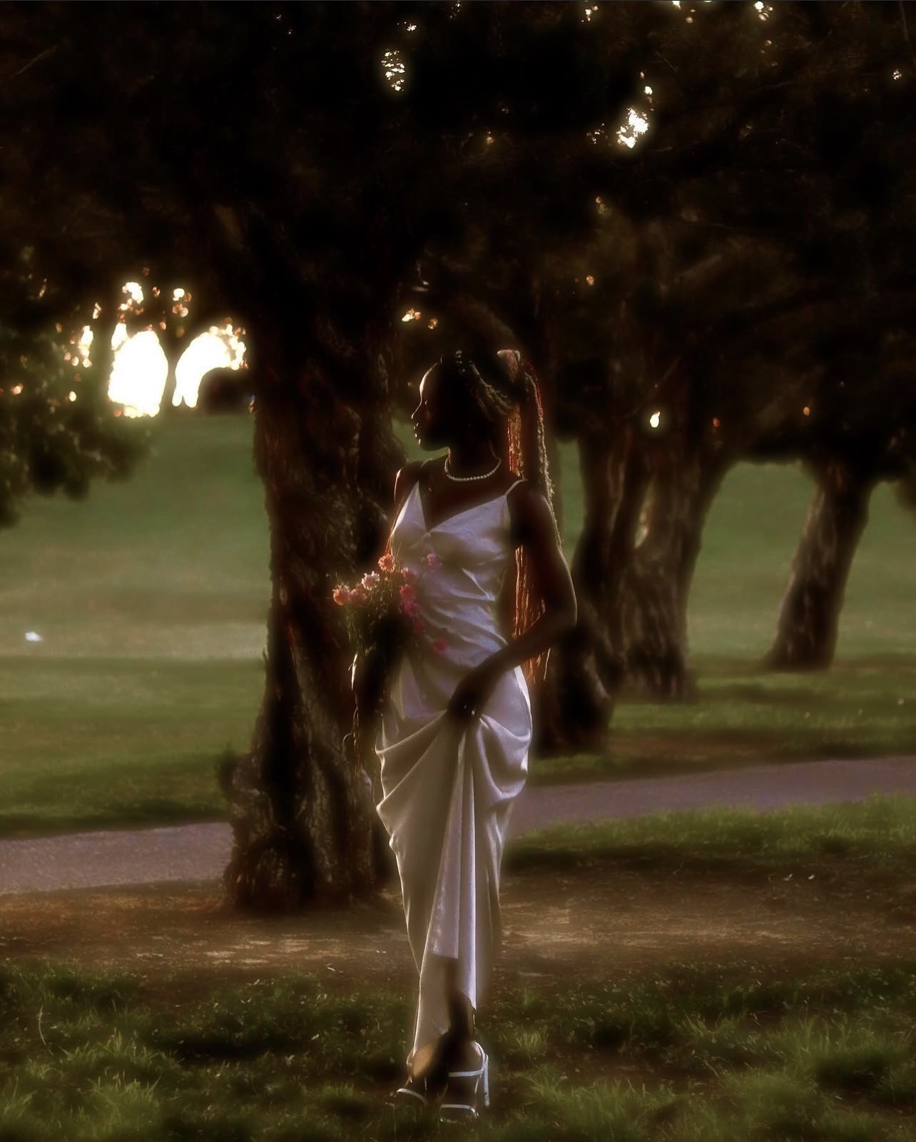Person in a white dress standing near a tree in a park during sunset, holding a flower, with trees and grass in background.