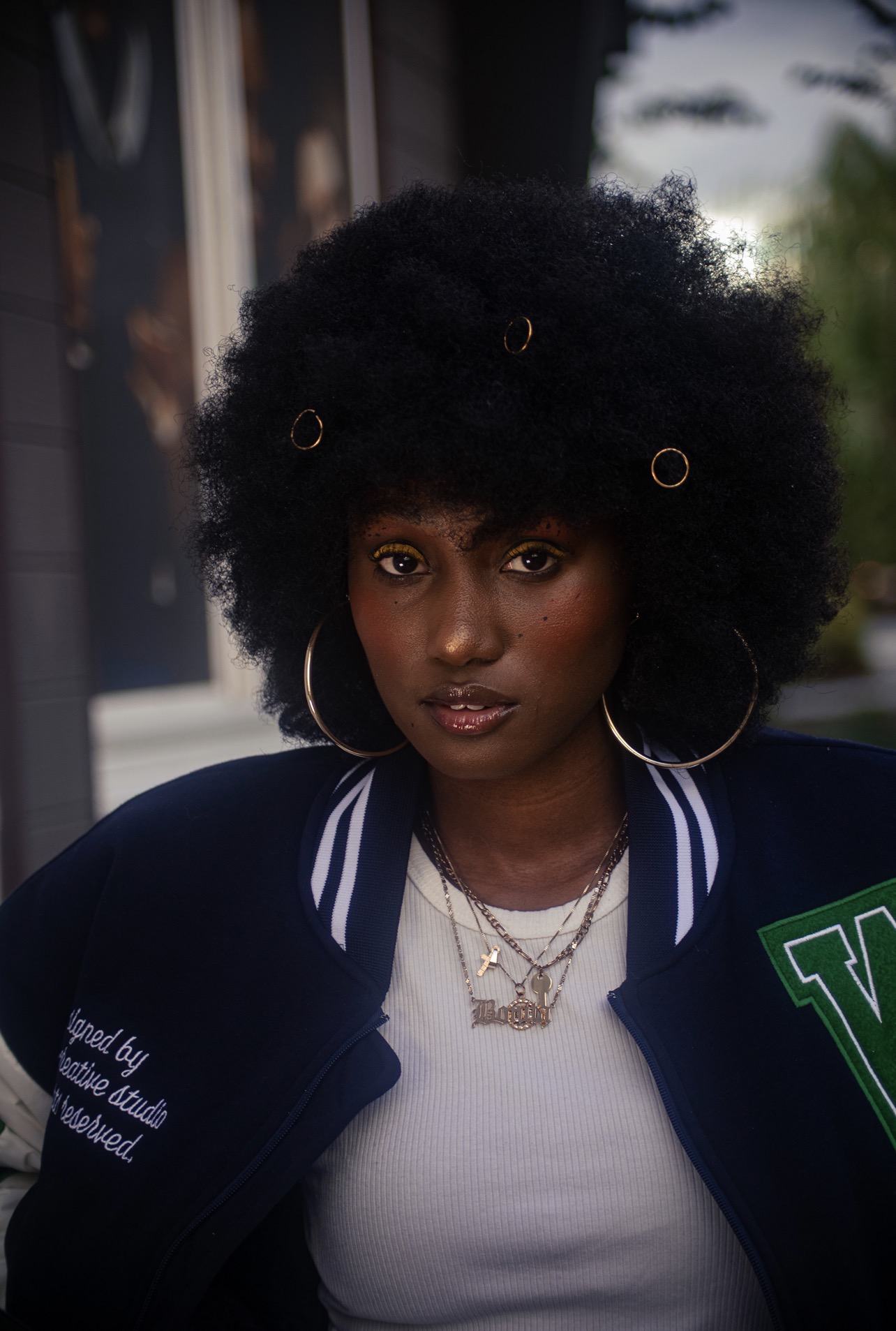 Woman with large curly hair wearing a navy jacket and white shirt, outdoors, with trees and building in background.