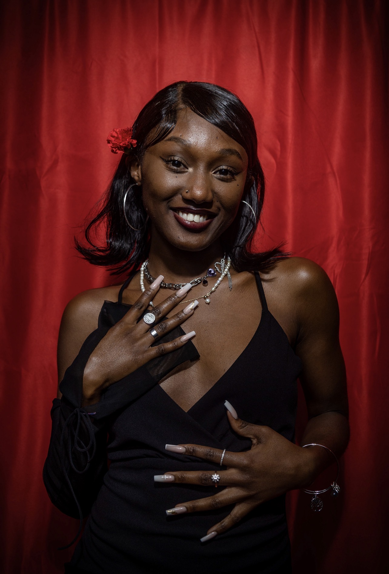 Smiling woman with dark hair and jewelry poses in front of red curtain, wearing a black dress.