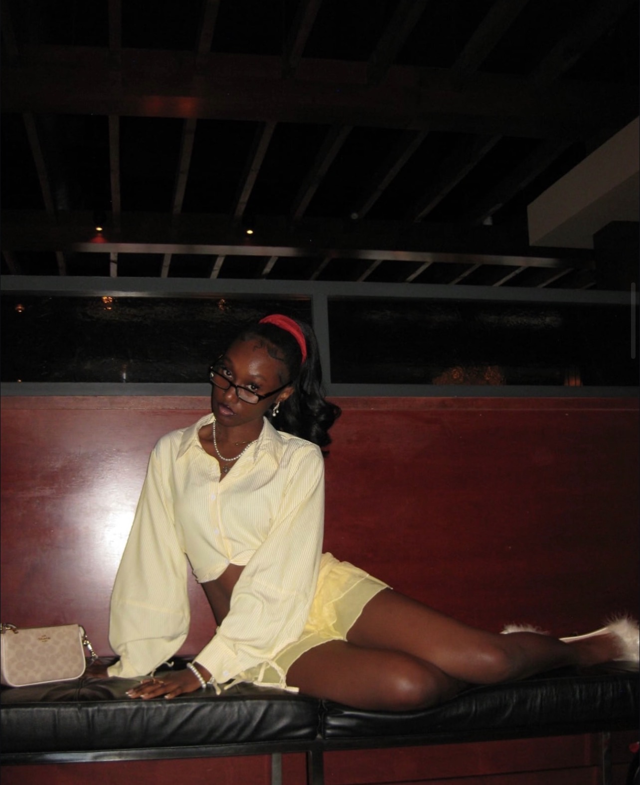 Person with glasses and a red headband sitting on a black surface, wearing a yellow outfit, in a dimly lit room.