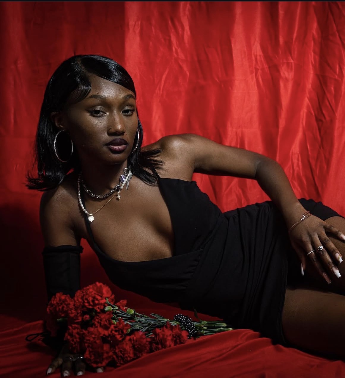 Woman in black dress lying on red fabric with red flowers, red curtain background, jewelry, and dark lipstick.
