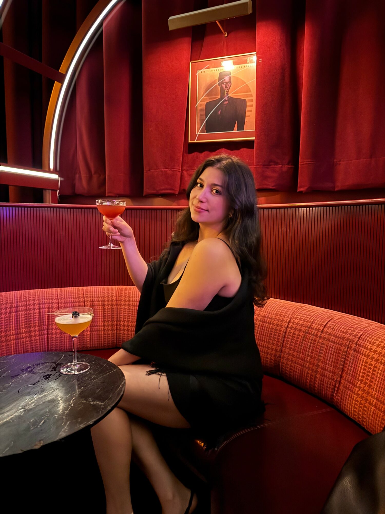 Woman with long dark hair sitting in a red booth holding a cocktail, with a framed portrait on red curtains behind her.