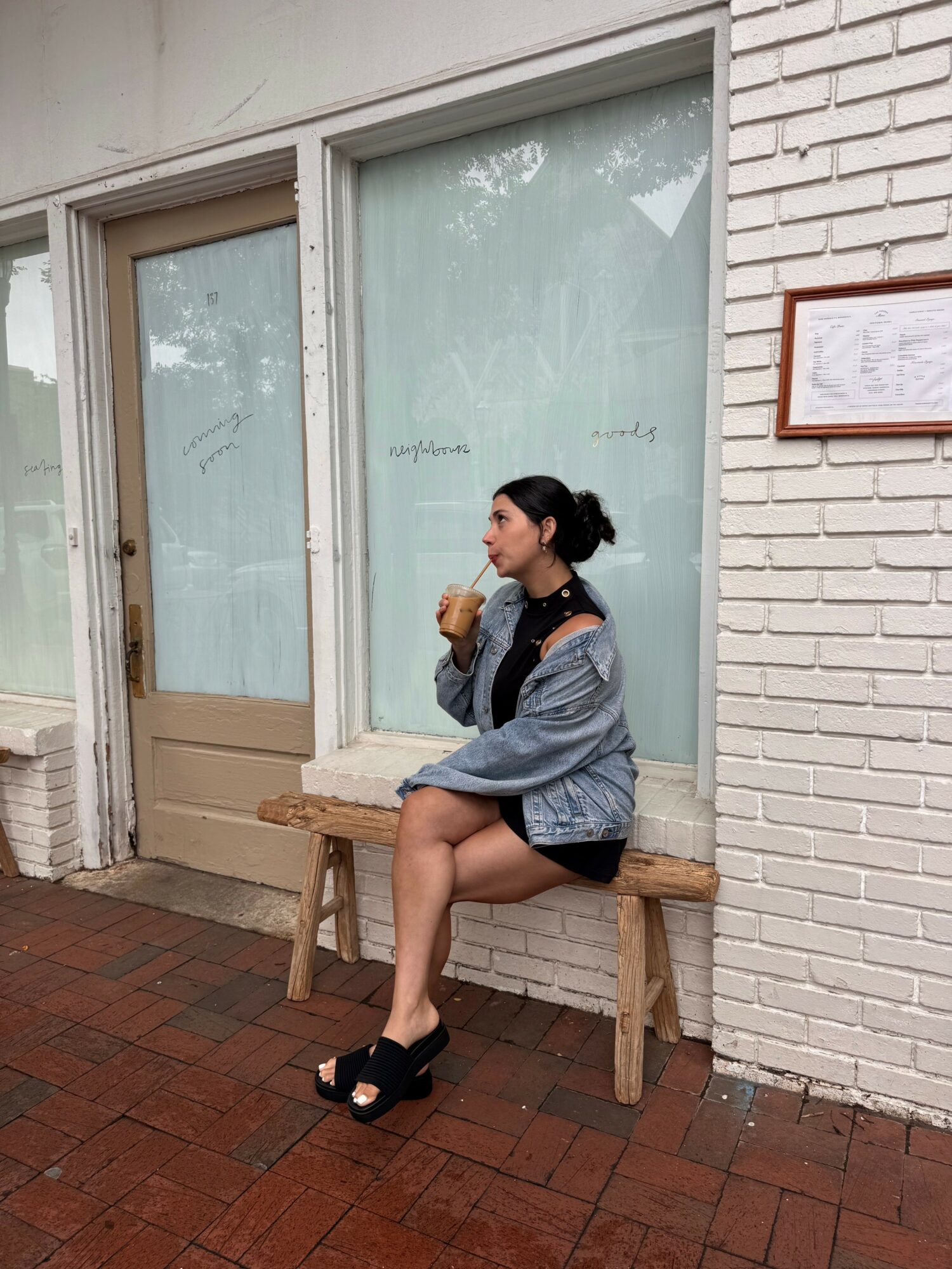 Young woman sitting on a wooden bench outside a building with large windows and a white brick wall, holding a drink.