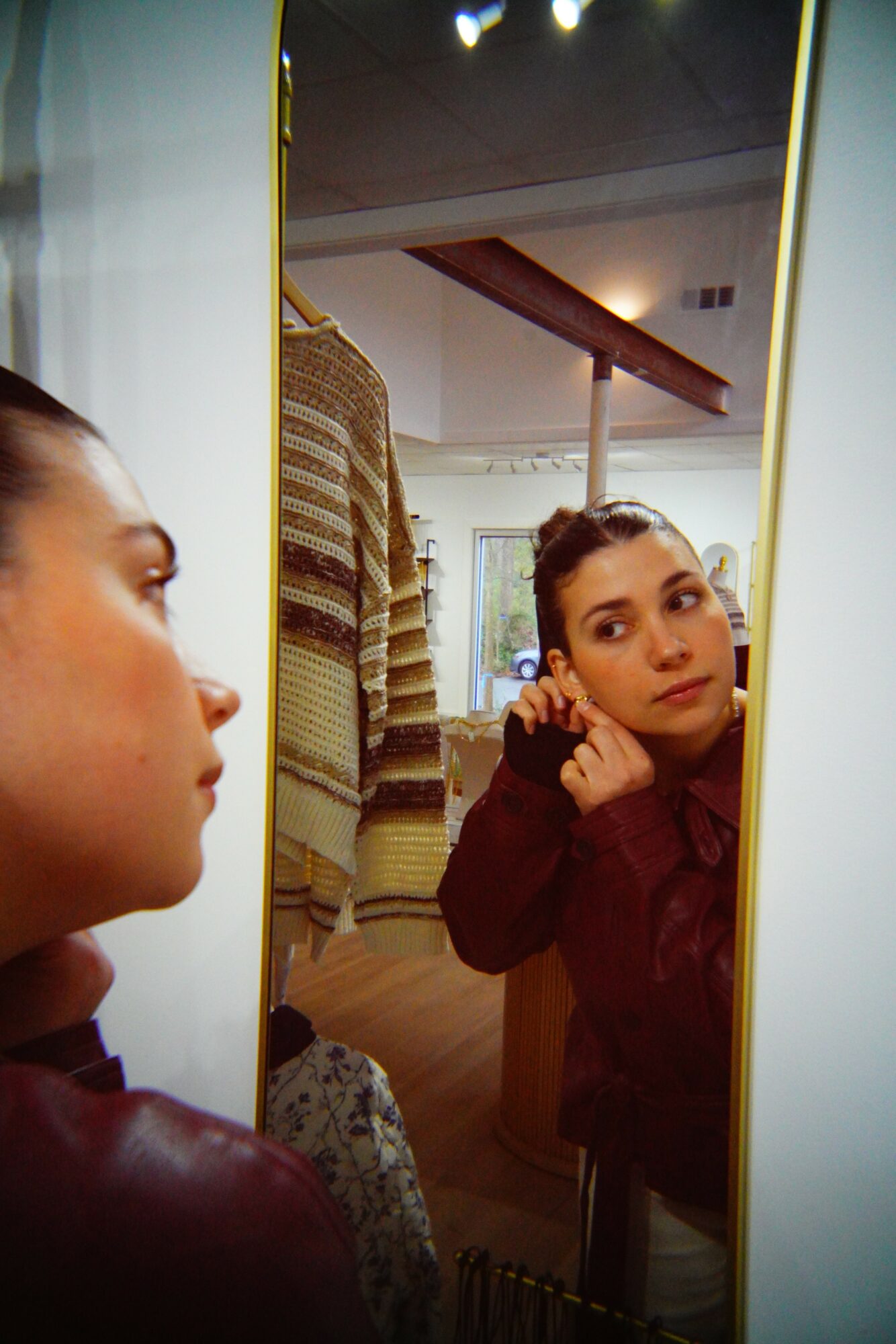 Young woman looking at her reflection in a mirror, adjusting her earring, with a clothing rack in the background.