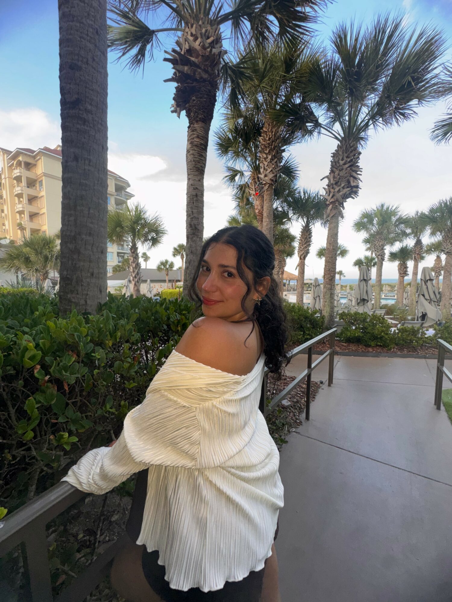 Woman with dark hair in a white off-shoulder top standing outdoors near palm trees and greenery.