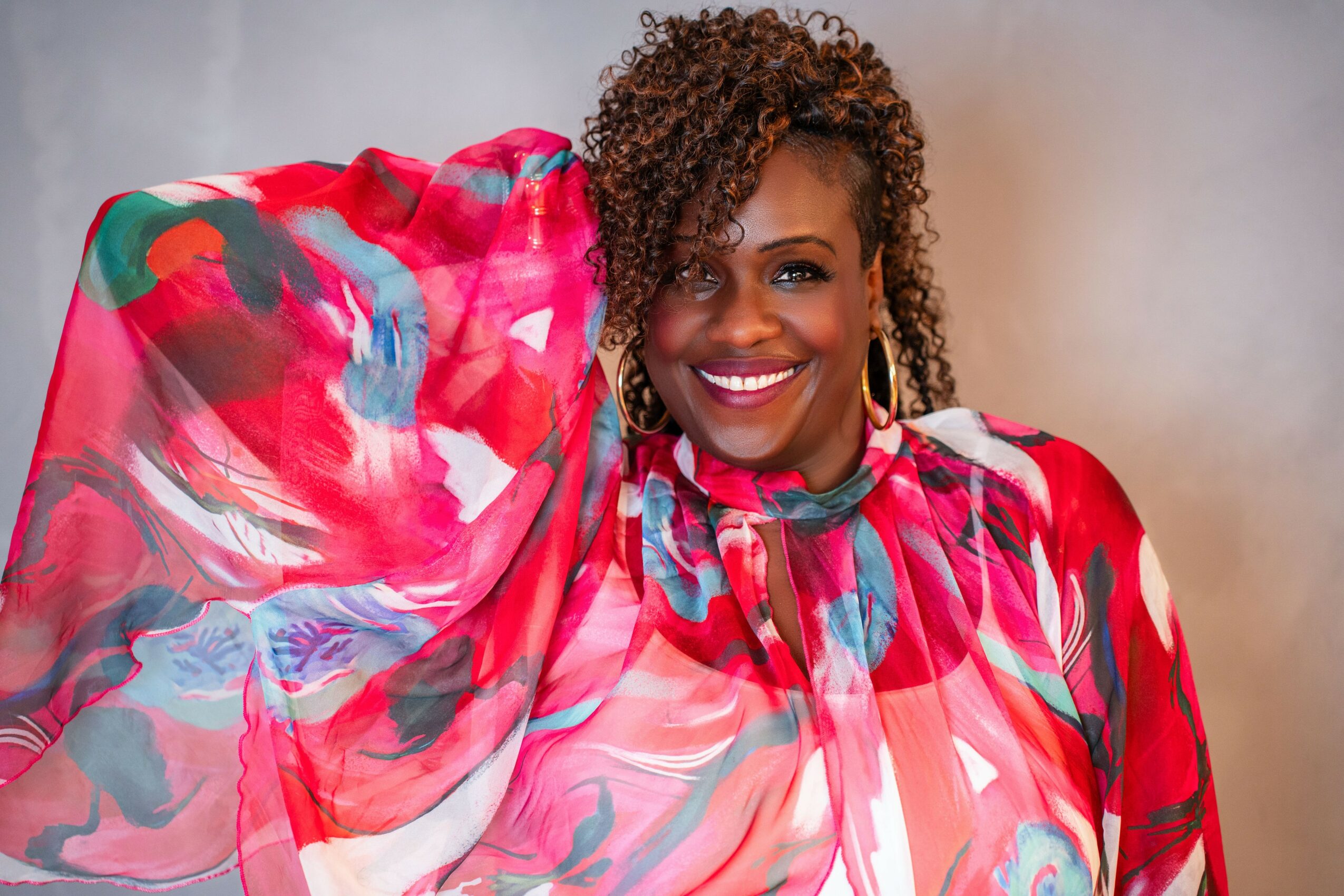 Smiling woman with curly hair wearing a colorful, patterned blouse, posing with her arm raised, against a neutral background.
