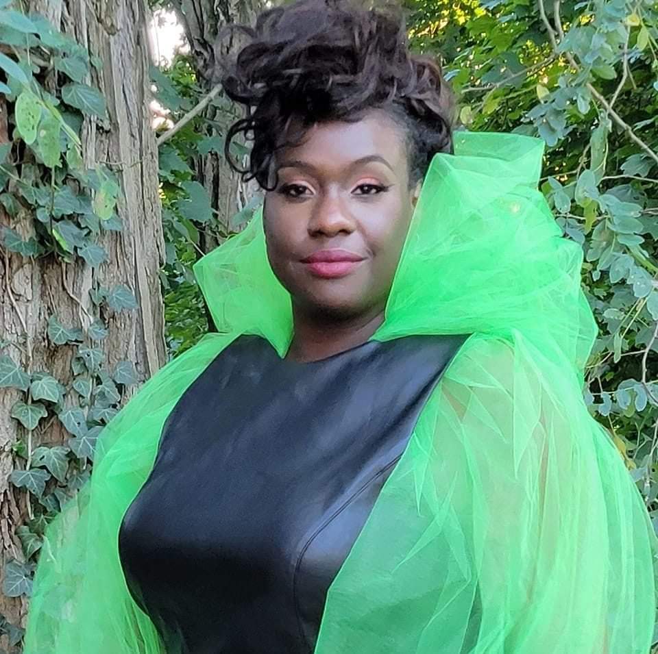 Woman with short curly hair wearing a black top and bright green sheer fabric, standing outdoors among green foliage.