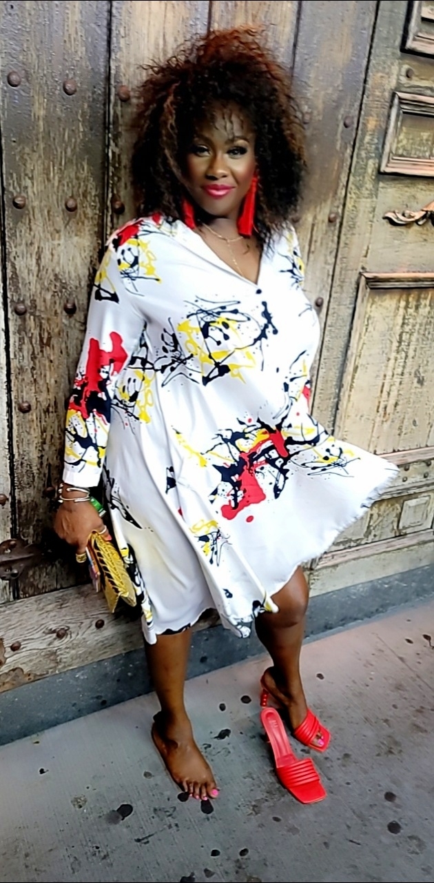 Woman with curly hair in a graffiti-style dress, standing barefoot with one red shoe, against a wooden wall.
