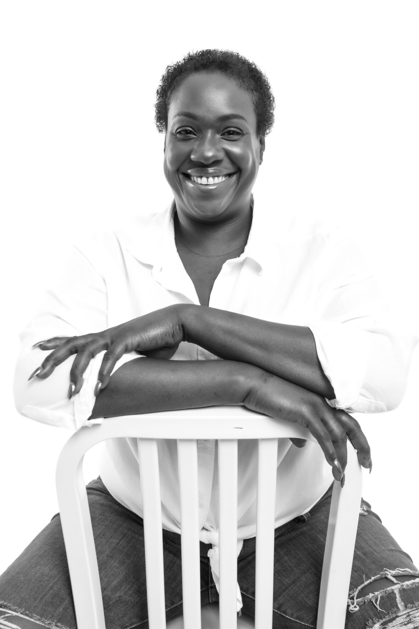 Smiling woman with short curly hair, sitting on a chair, arms crossed, wearing a white shirt, against a white background.