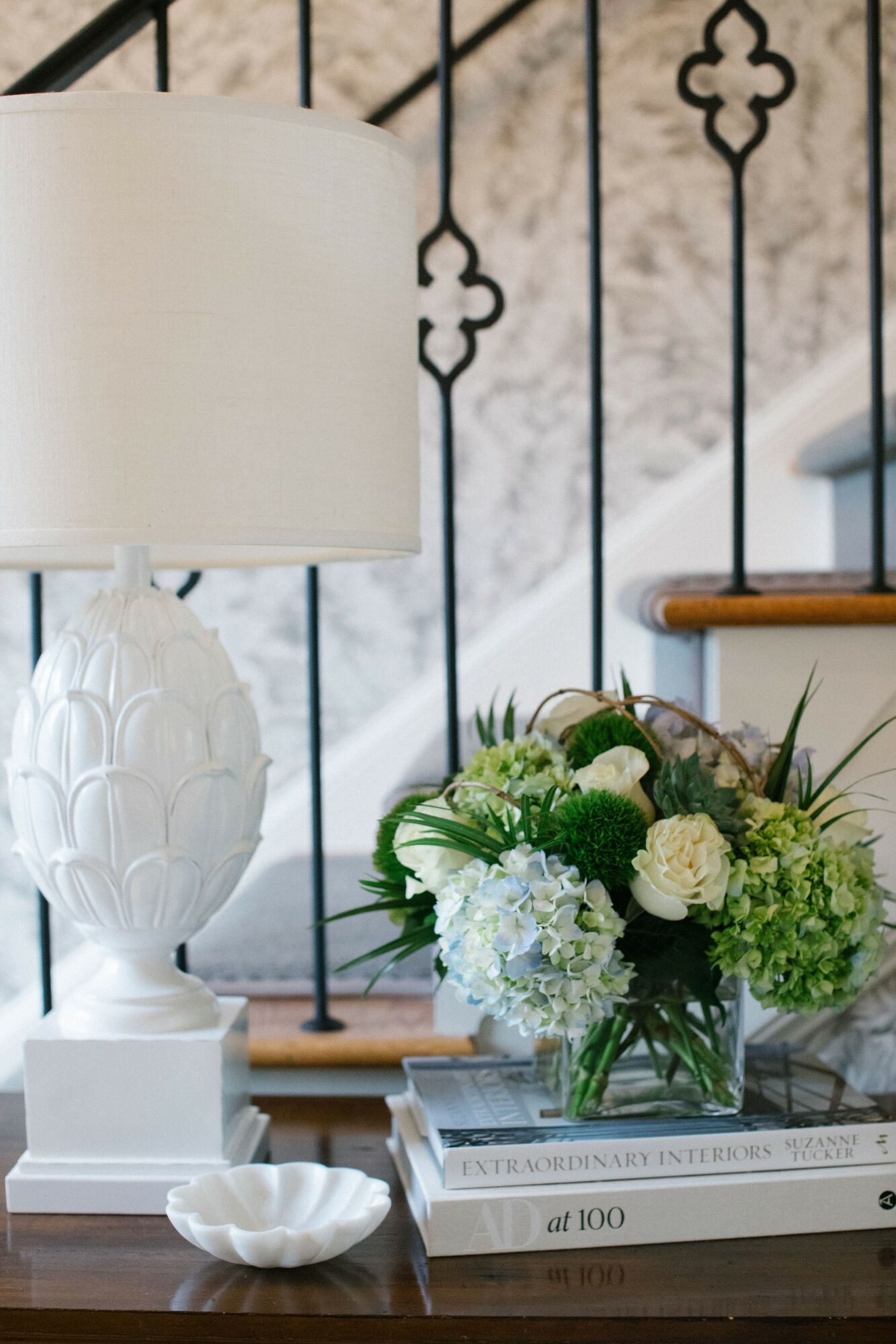 Table with a white lamp, a flower arrangement, and books, against a decorative black metal railing background.