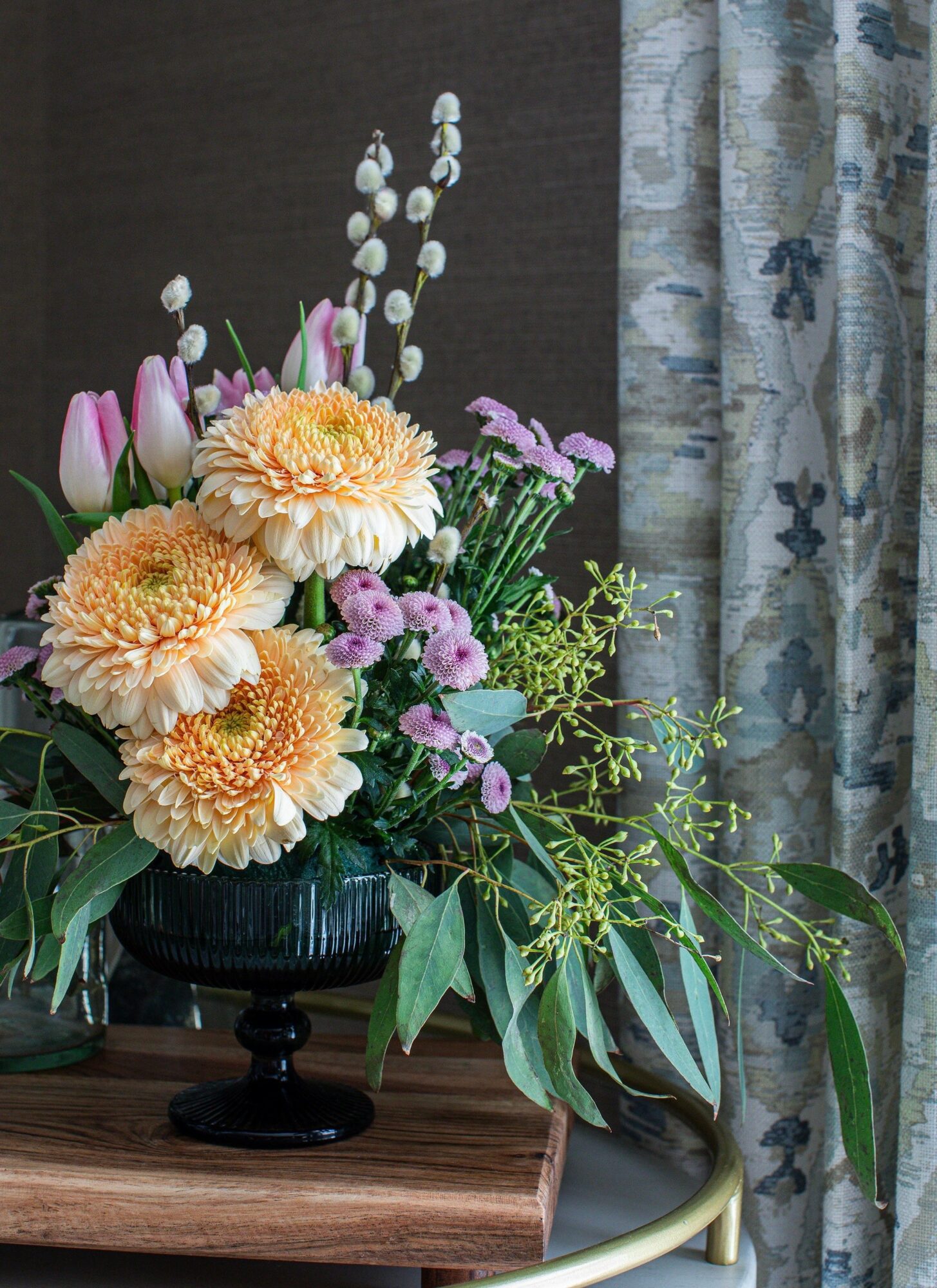 Flower arrangement with pink, yellow, and purple flowers in a black vase on a wooden surface, next to a patterned curtain.