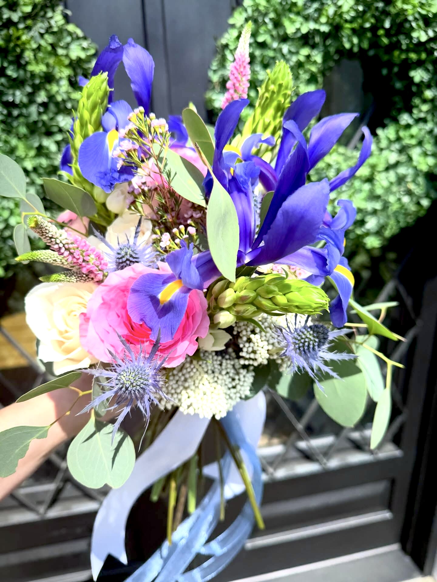 Colorful flower bouquet with purple, pink, and white flowers, green leaves, and a light blue ribbon, outdoors.