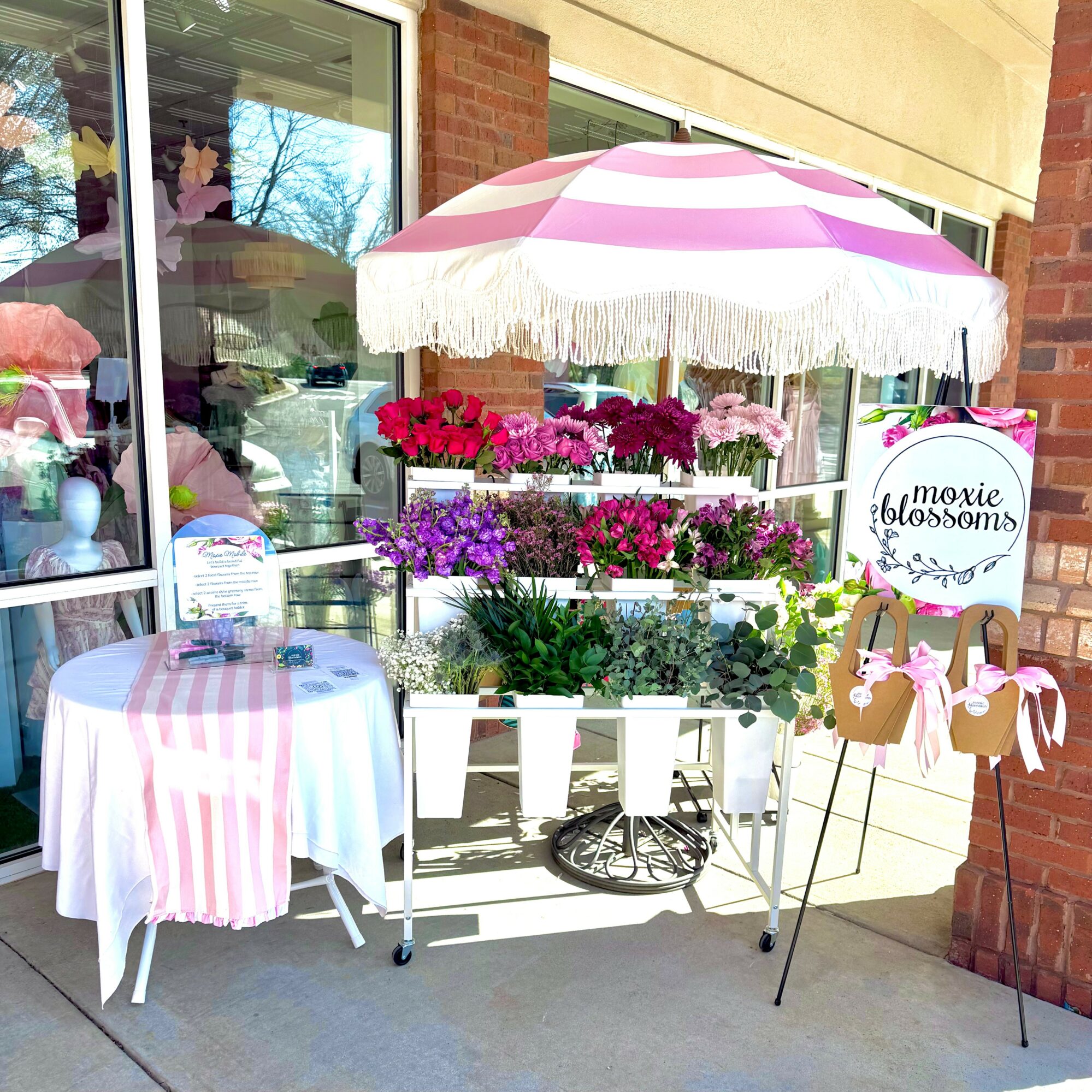 Flower stand with pink, purple, and white blossoms under a pink and white striped umbrella outside a shop.