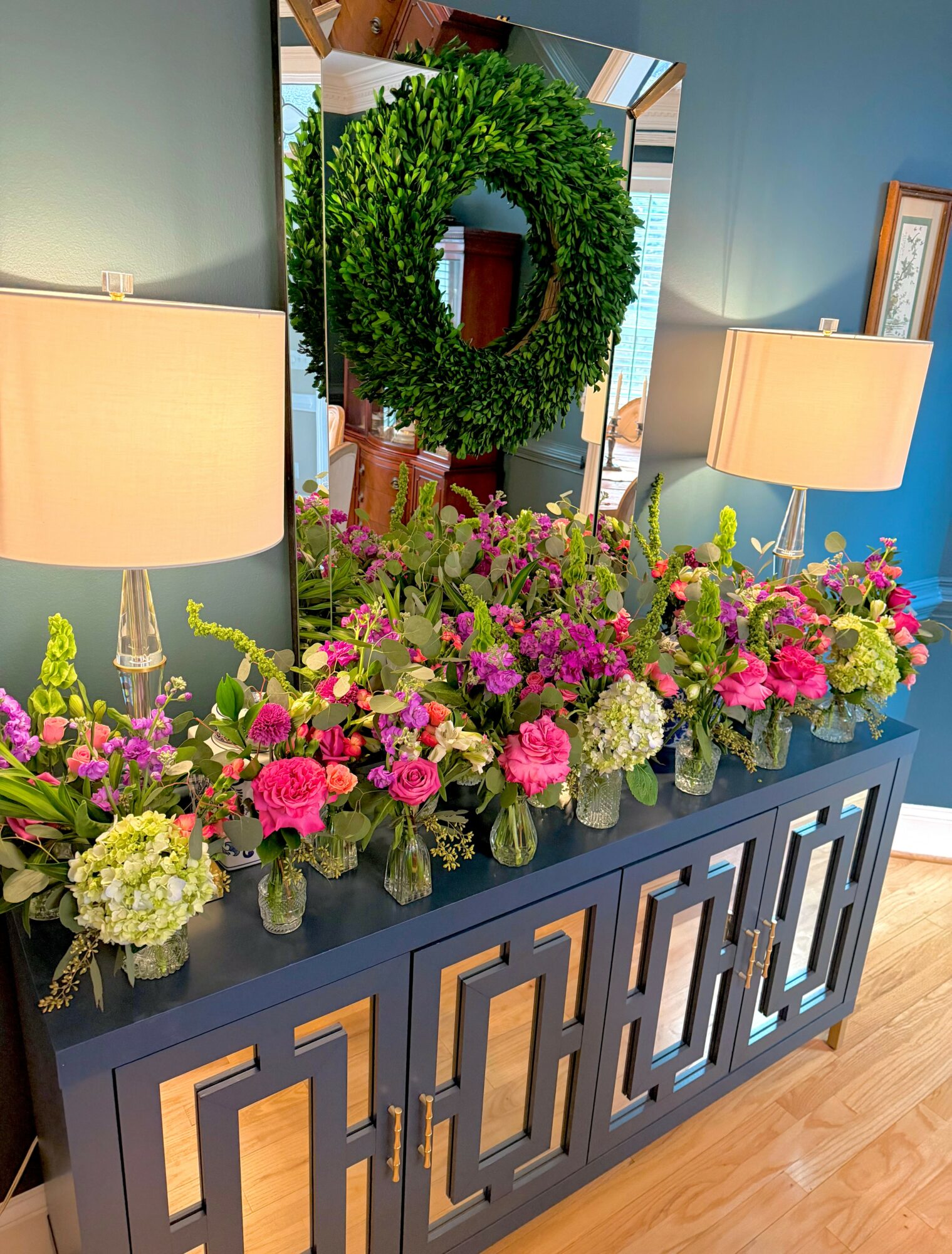 Decorative blue sideboard with pink, purple, and white flowers, two lamps, and a mirror with a green wreath reflection.