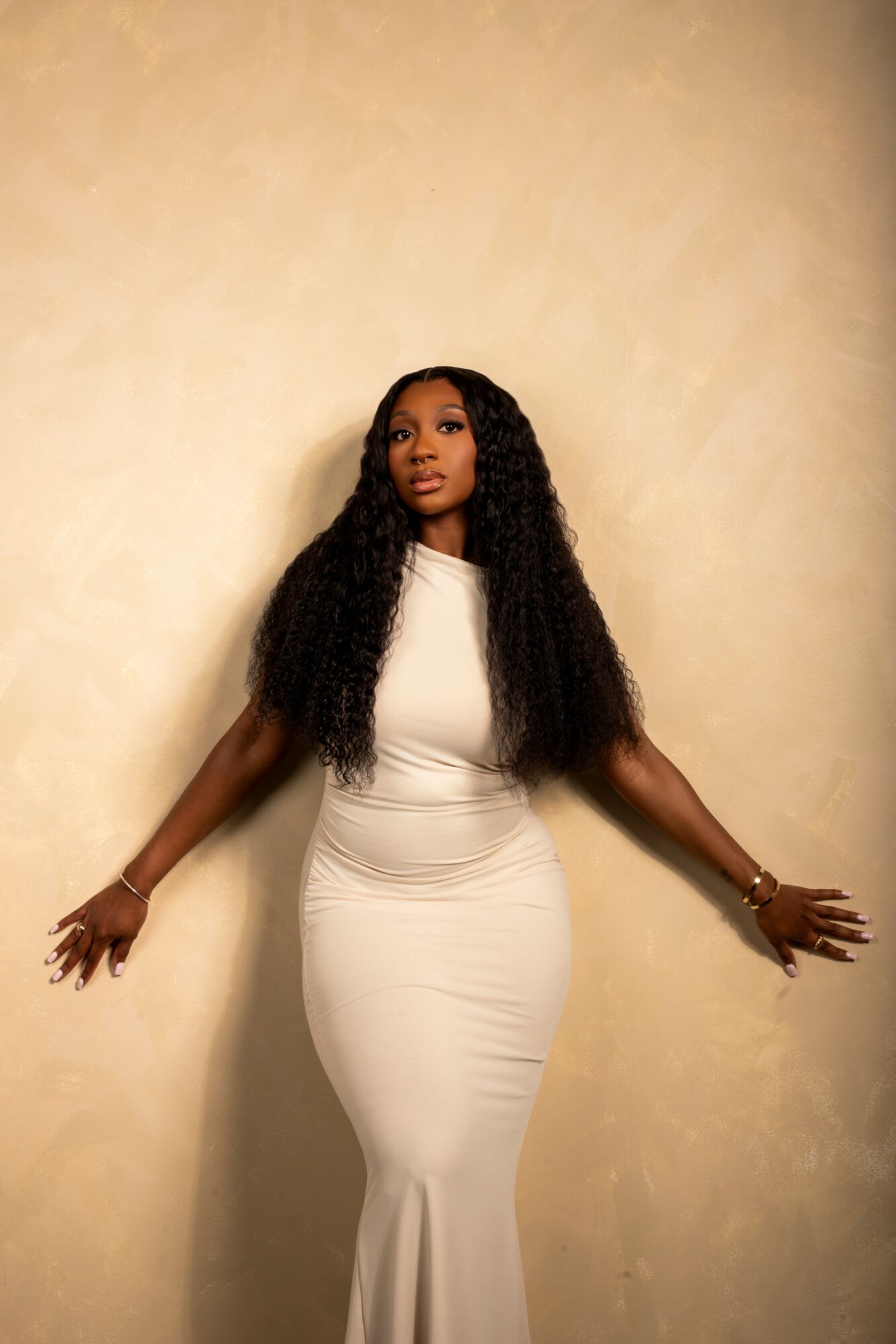 Woman in a white dress standing against a beige wall with arms outstretched, looking upward.