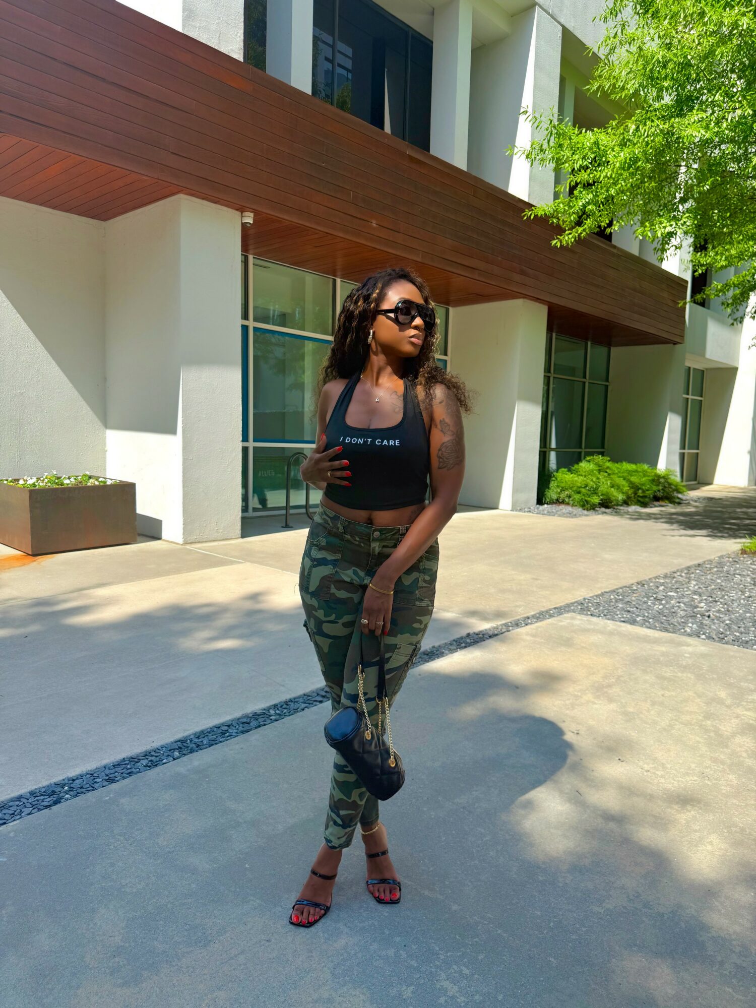 Woman with sunglasses standing outdoors in front of modern building, wearing a black top, camouflage pants, and heels.