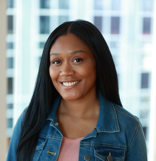 Smiling woman with long black hair wearing a denim jacket and pink top, standing indoors with a cityscape background.
