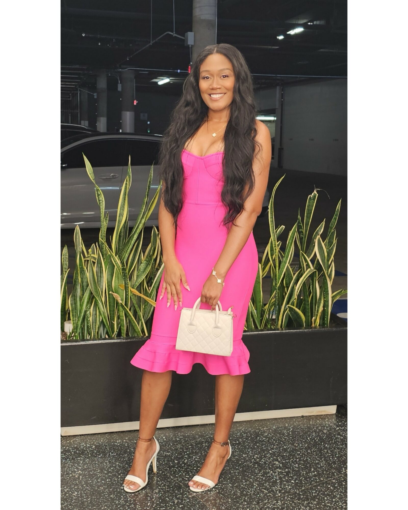 Woman in pink dress holding a white handbag, standing outdoors with plants and a parking garage in background.