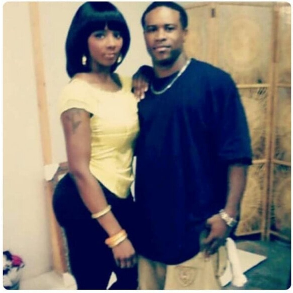 A woman and a man stand together indoors, posing for a photo. The woman wears a yellow top, and the man wears a dark shirt.