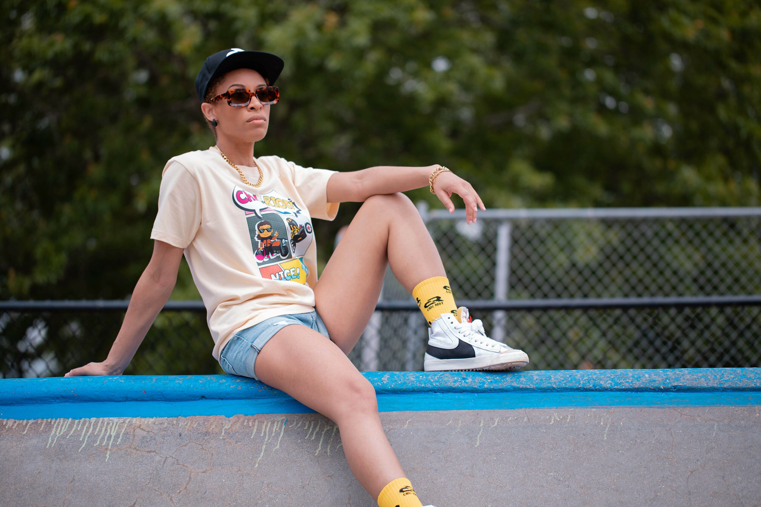 Person sitting on a ledge outdoors, wearing sunglasses, a cap, a graphic t-shirt, shorts, and sneakers, with trees in background.