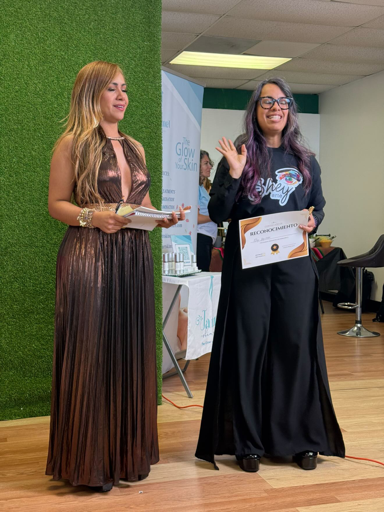 Two women standing indoors, one holding a certificate and the other waving, with a green wall and presentation setup behind them.