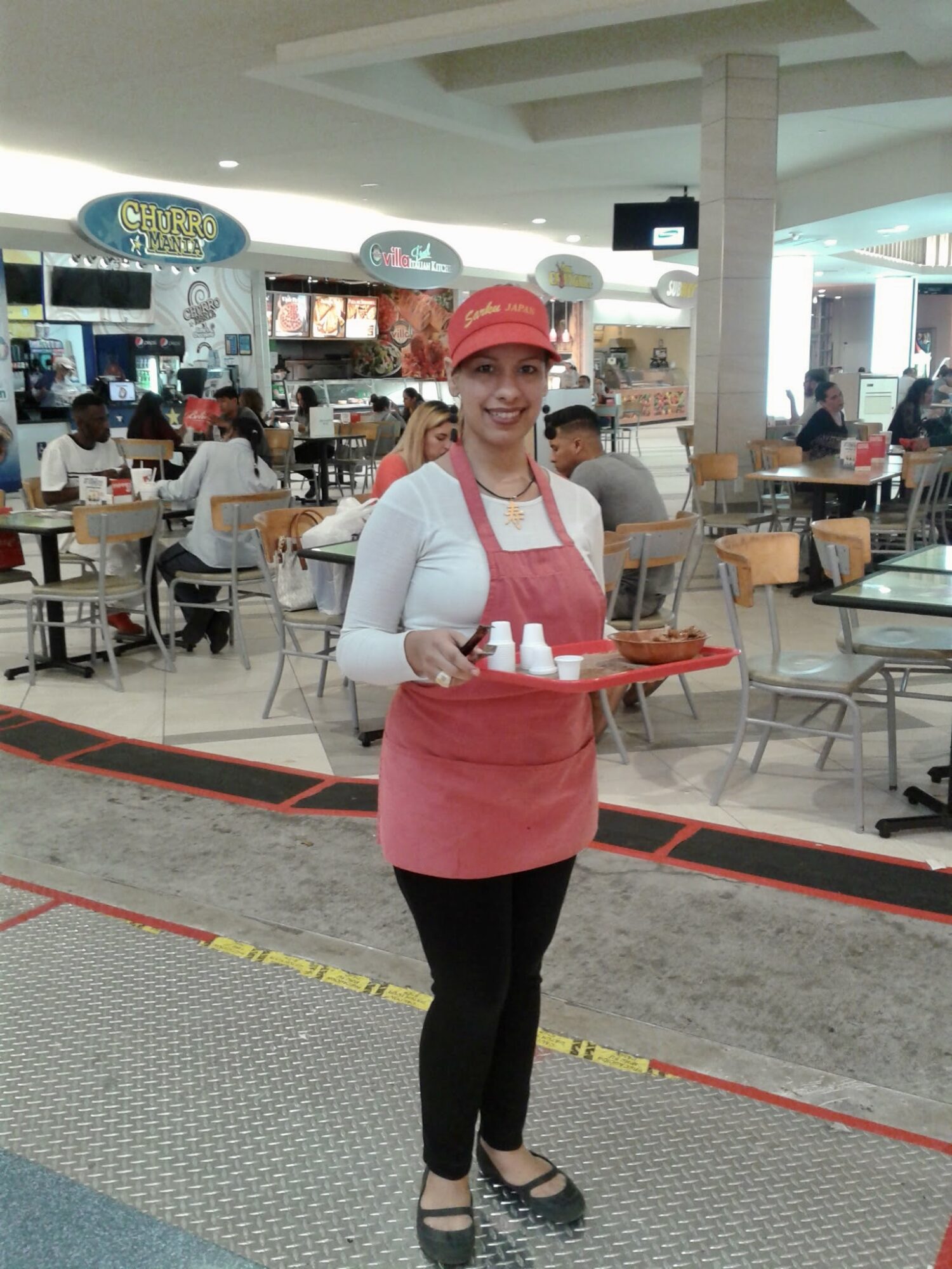 Woman in red apron and cap holding a tray with cups in a food court.
