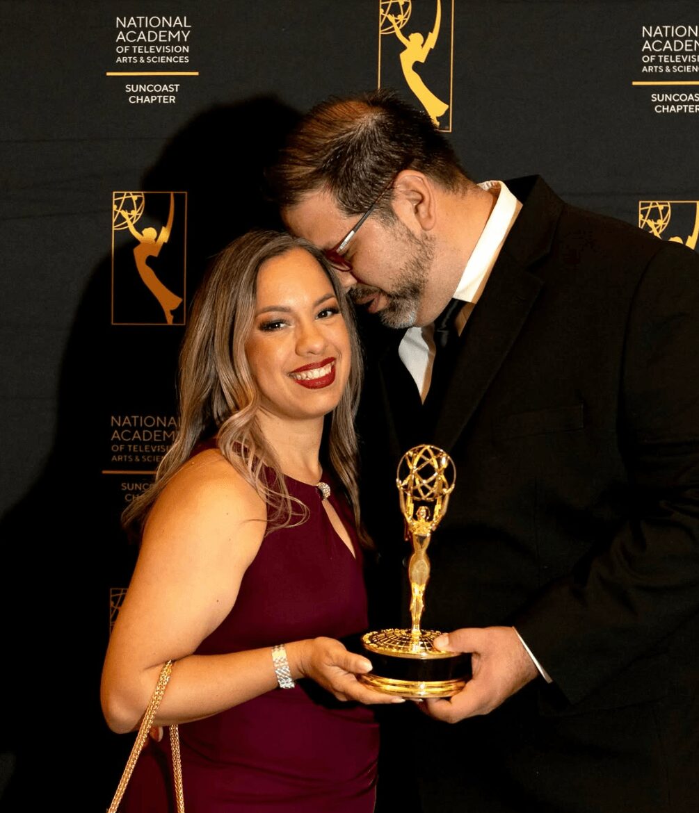 A woman and a man hold an Emmy award together, smiling, at an awards event backdrop.