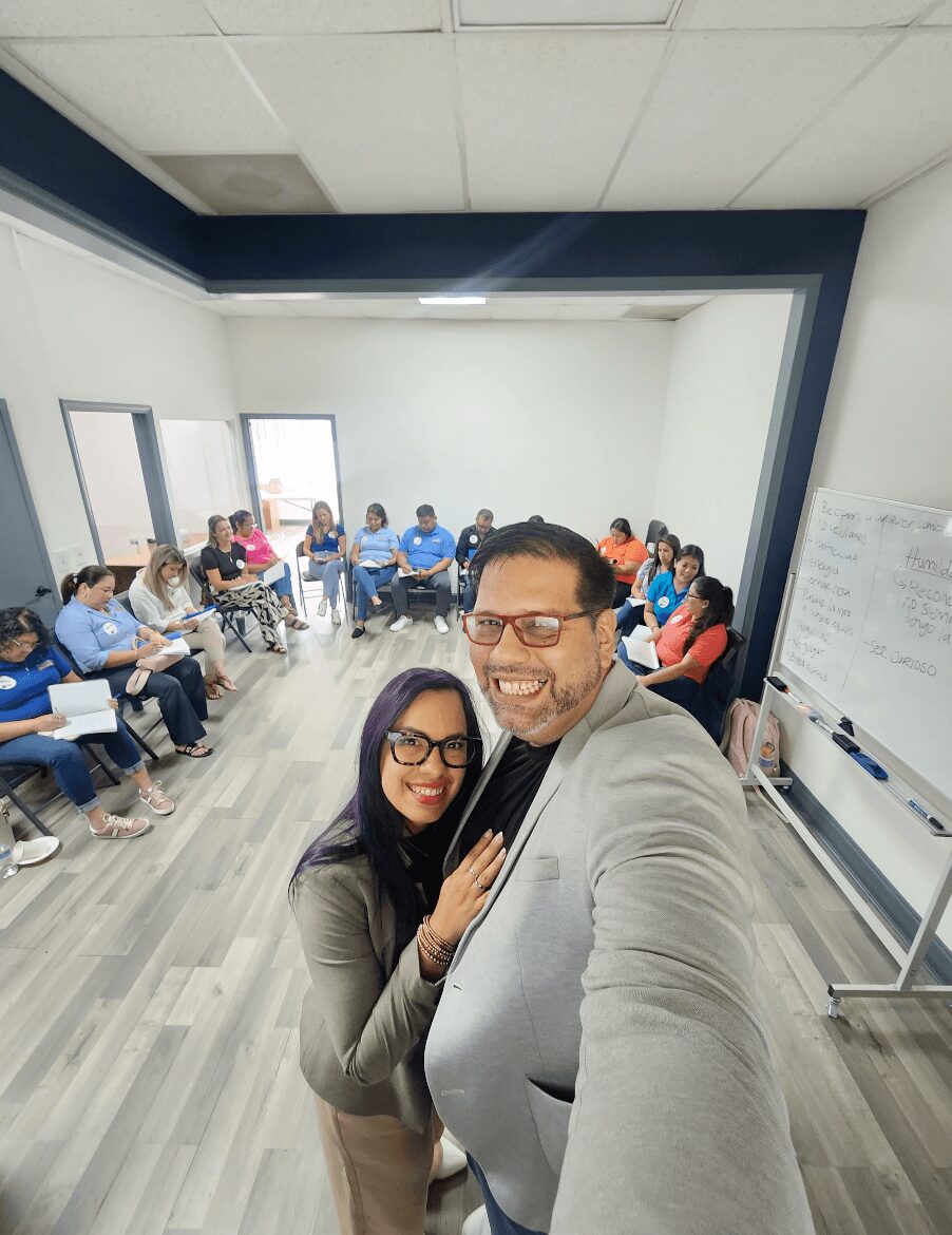 Two people taking a selfie in a classroom with students seated around the room, whiteboard on the wall, and windows in the background.