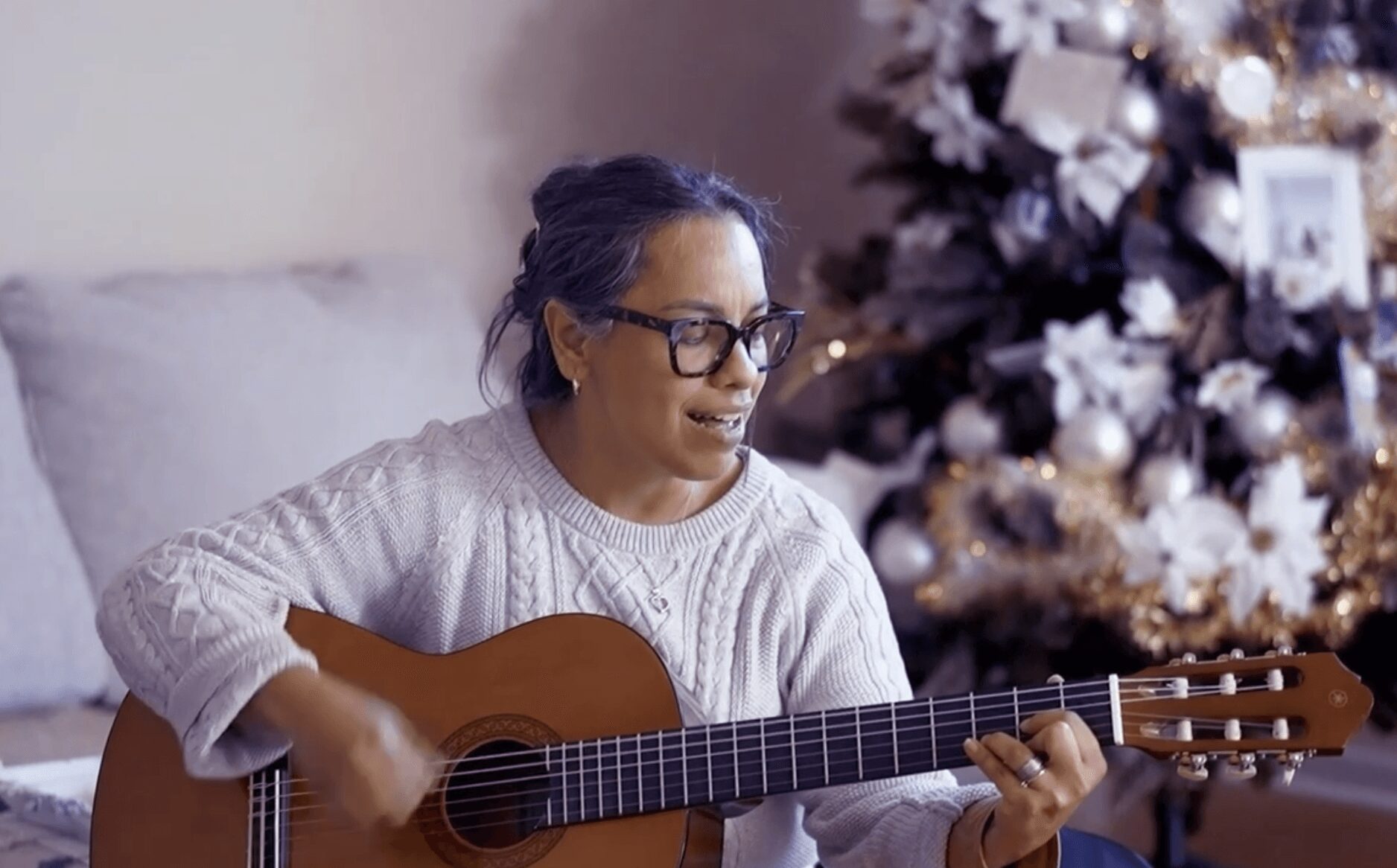 Woman with glasses playing guitar indoors near decorated Christmas tree, smiling, wearing a white sweater.